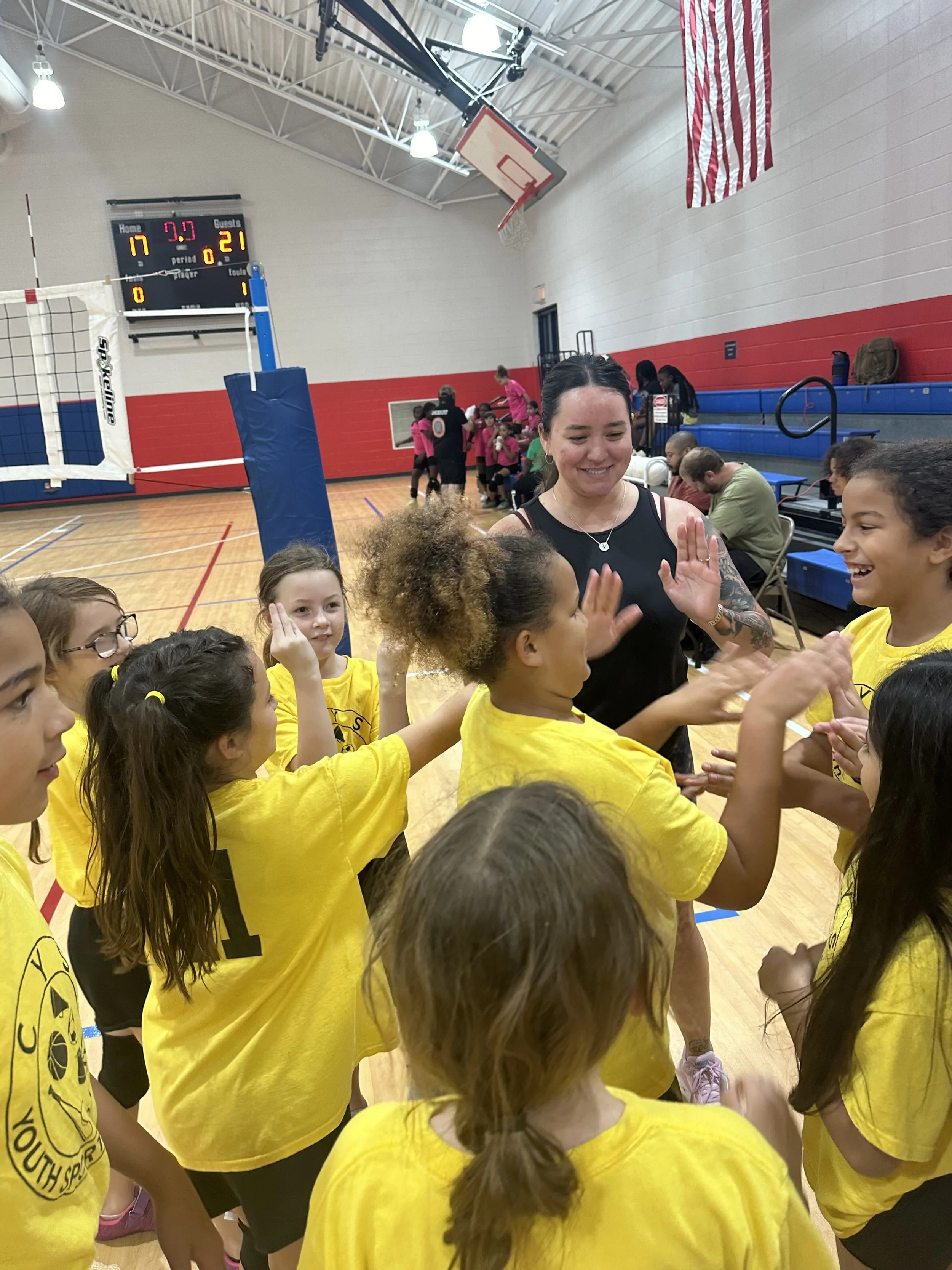 Picture of adult female wearing a black tank top high fiving various children wearing yellow shirts after a volleyball game for 10 and under youth.