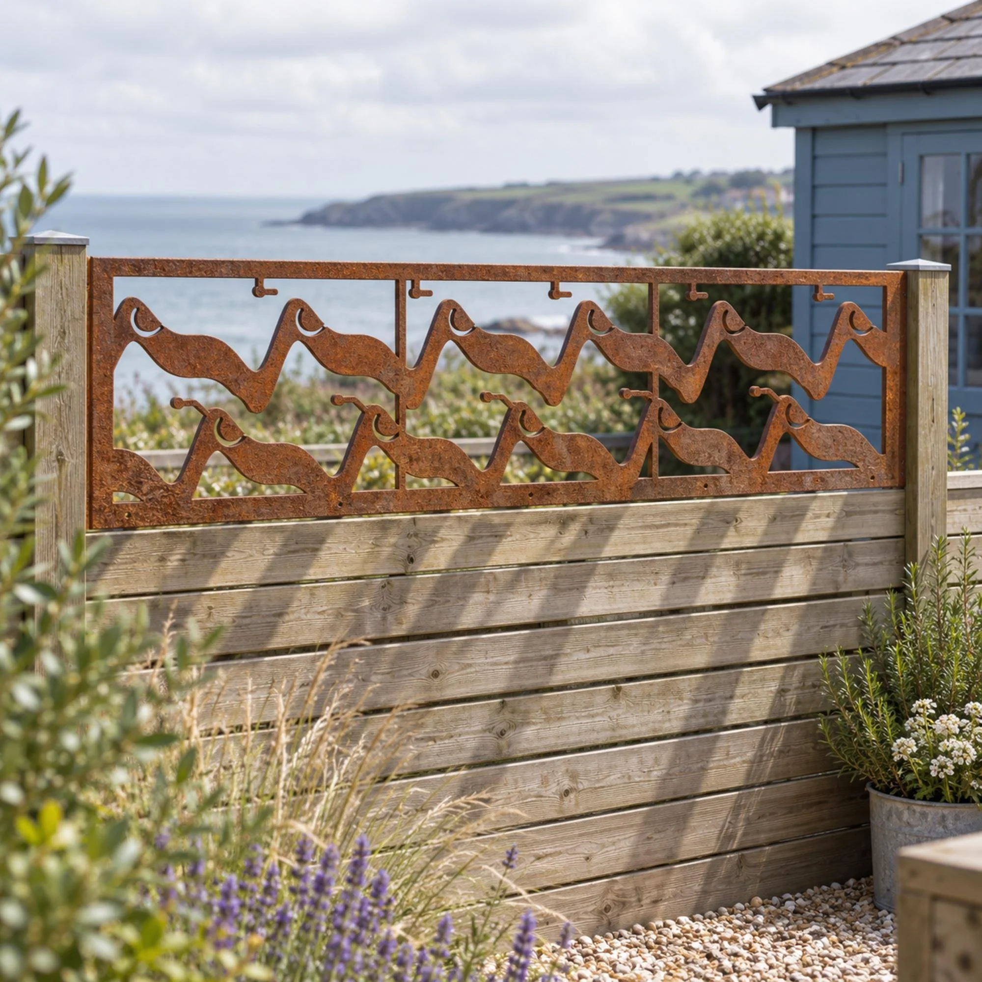 Tidewater Fence Topper in weathered raw steel, fixed along a 1.8 m horizontal-slat timber fence in a small seaside terrace garden with a blue-painted shed and gravel