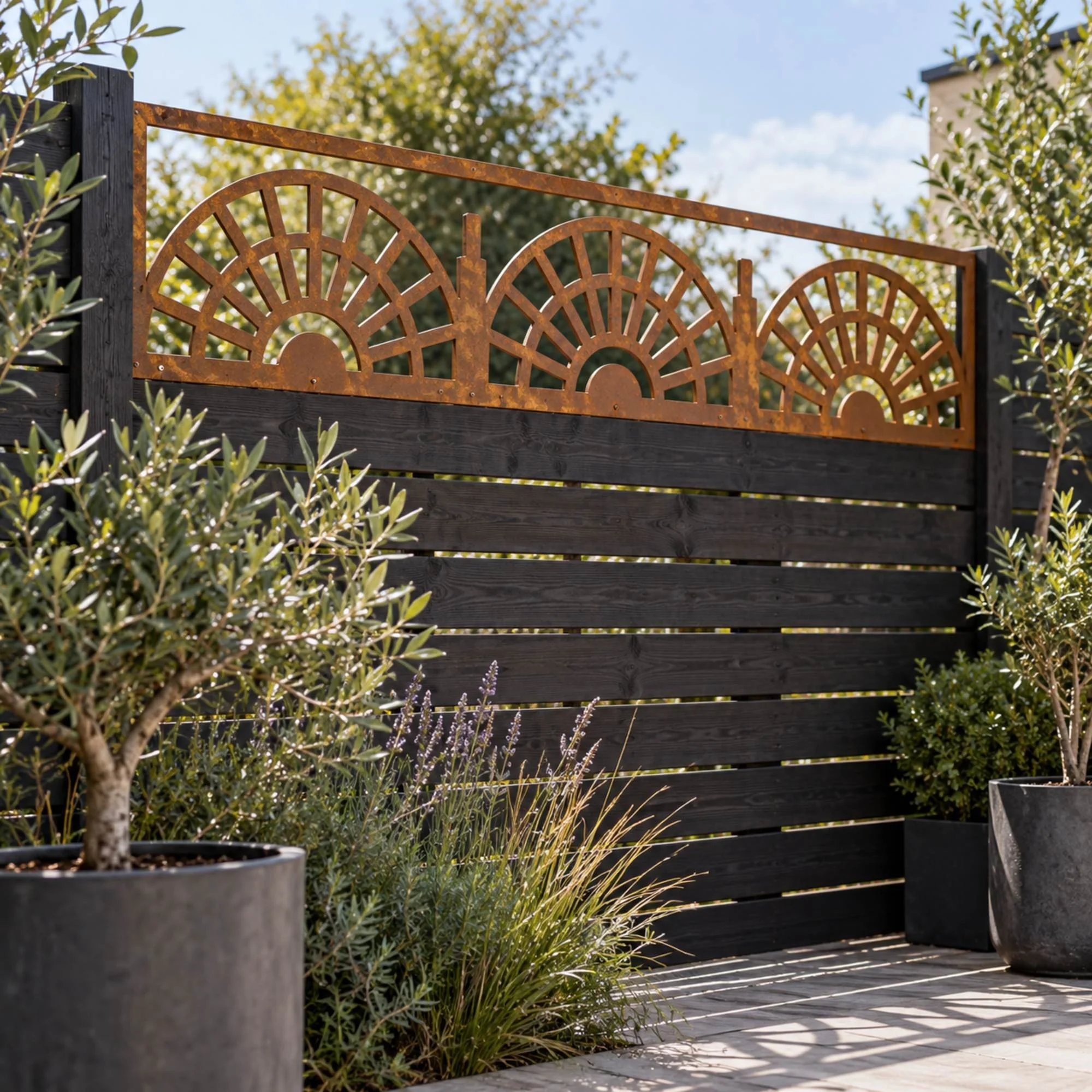 Ember Fence Topper in weathered raw steel, fixed along a 1.8 m black-stained horizontal-slat fence in a compact modern courtyard with grey porcelain paving and potted olive trees