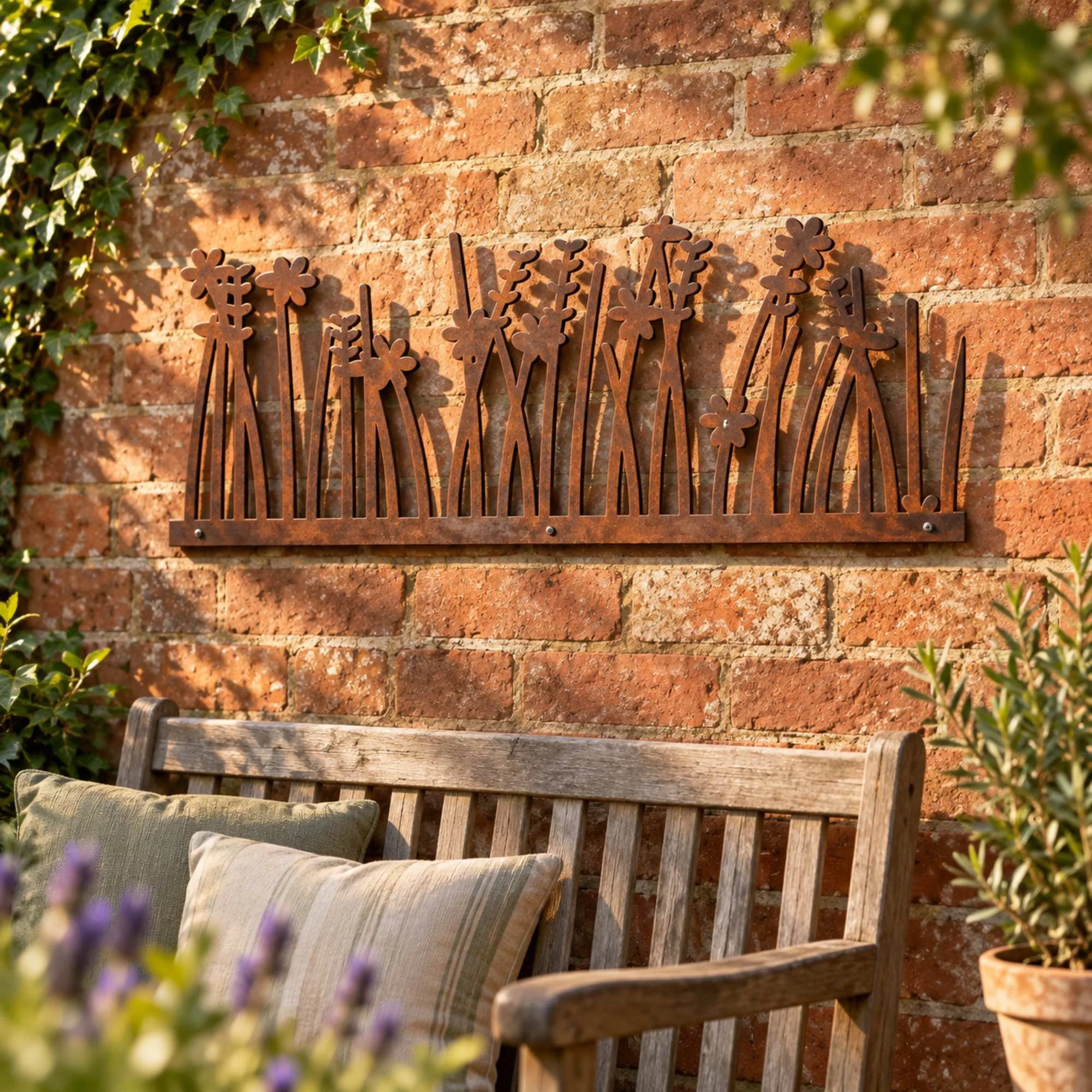 Saltmarsh Meadow Metal Wall Art in weathered raw steel, fixed to a red brick courtyard wall above a two-seat wooden garden bench in a UK back garden