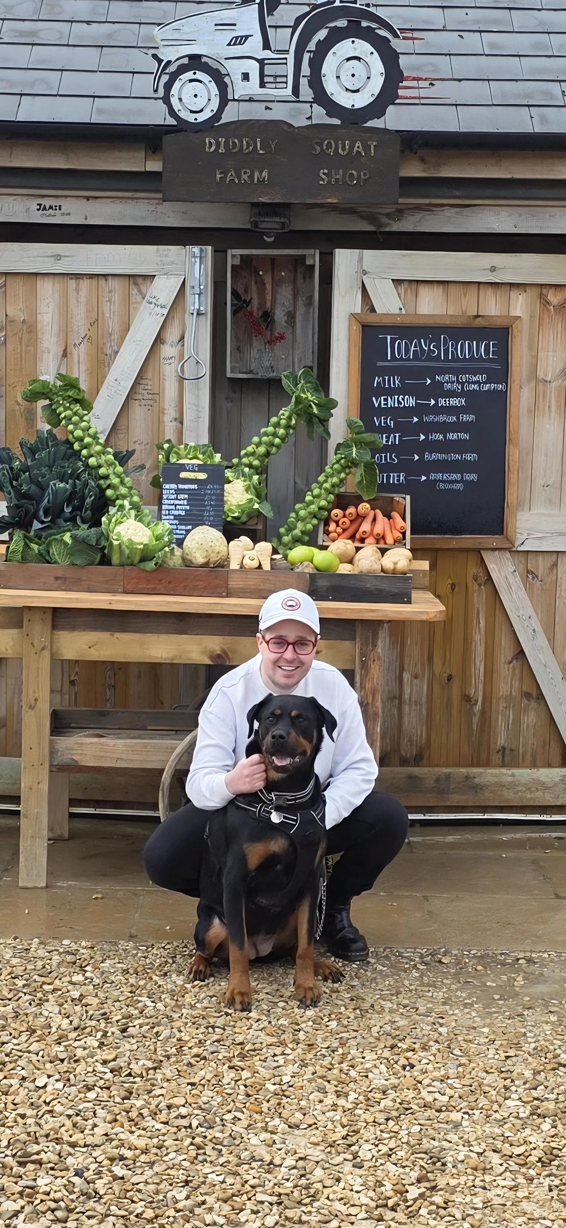 A man in a white hoodie and red sunglasses kneeling next to a black and brown dog in front of a vegetable stand at a farm shop. The stand has fresh produce like carrots, potatoes, and greens, and a chalkboard sign listing today's produce. There is a sign with a tractor illustration above the stand.