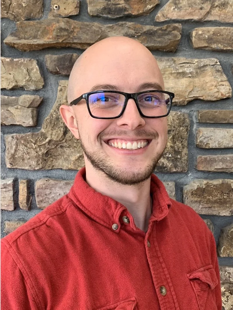 A man with glasses, a beard, and a big smile wearing a red shirt, standing in front of a stone wall.
