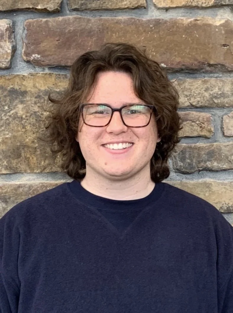 A smiling person with curly brown hair, glasses, and a navy blue shirt standing in front of a brick wall.