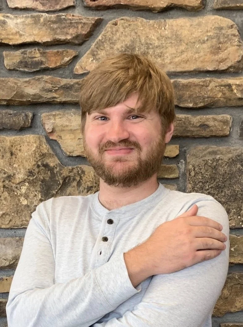 A man with brown hair and a beard smiling with his arms crossed in front of a stone wall.