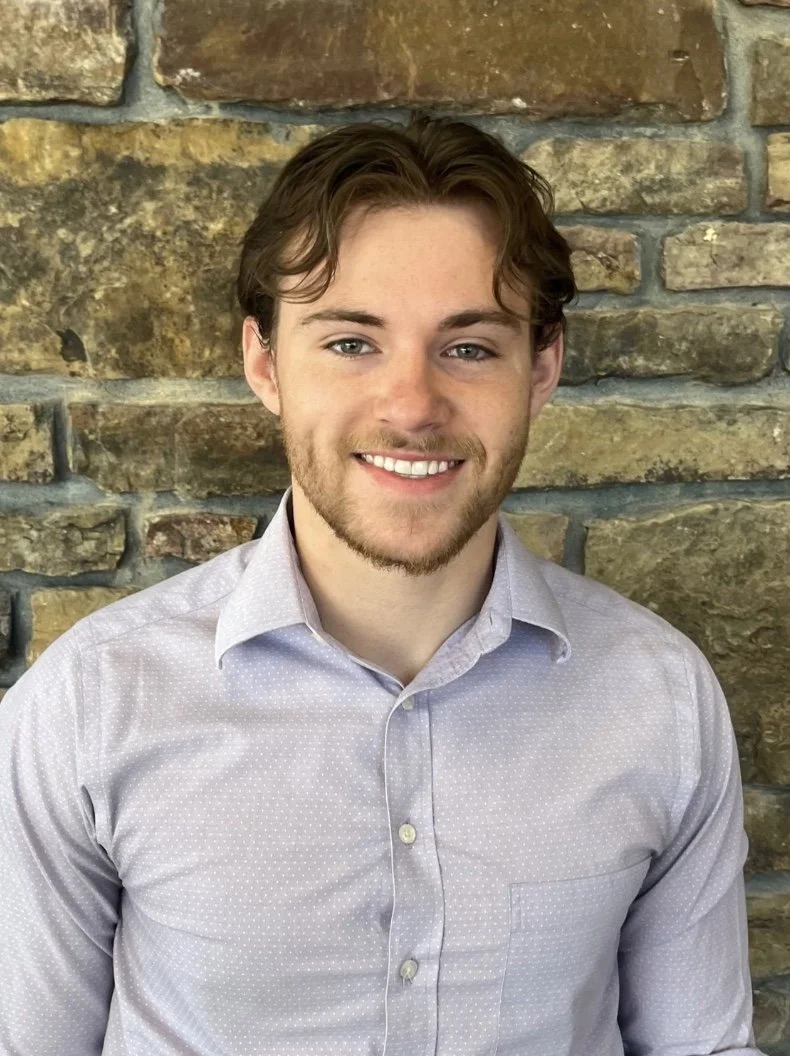 A young man with brown hair and a beard smiling, wearing a light purple button-up shirt, standing in front of a stone wall.