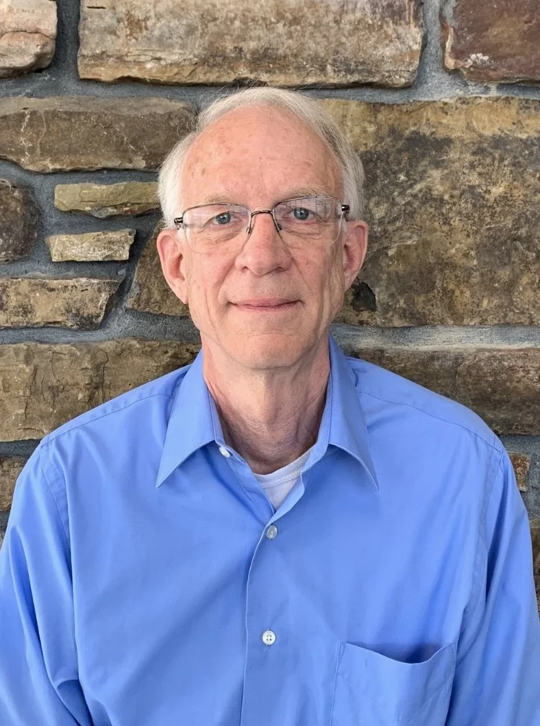 Portrait of an elderly man with glasses, wearing a light blue button-up shirt, sitting against a stone wall background.