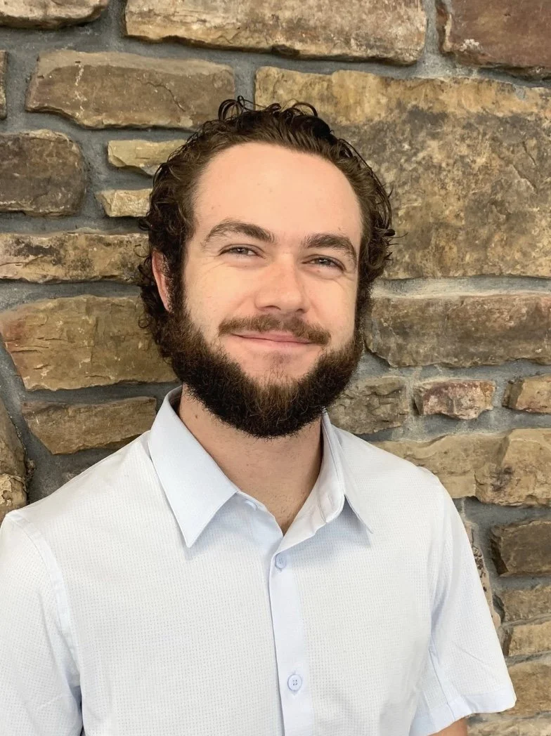 A young man with dark curly hair and a beard, wearing a white dress shirt, standing in front of a stone brick wall, smiling.