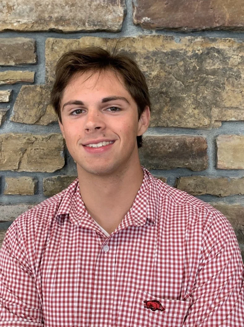 Young man with short brown hair, wearing a red and white checkered shirt, smiling, standing in front of a stone wall.