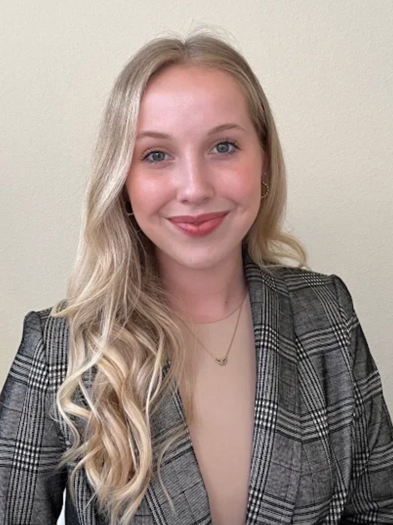 A young woman with long blonde wavy hair, wearing a plaid blazer and smiling at the camera against a plain beige wall.
