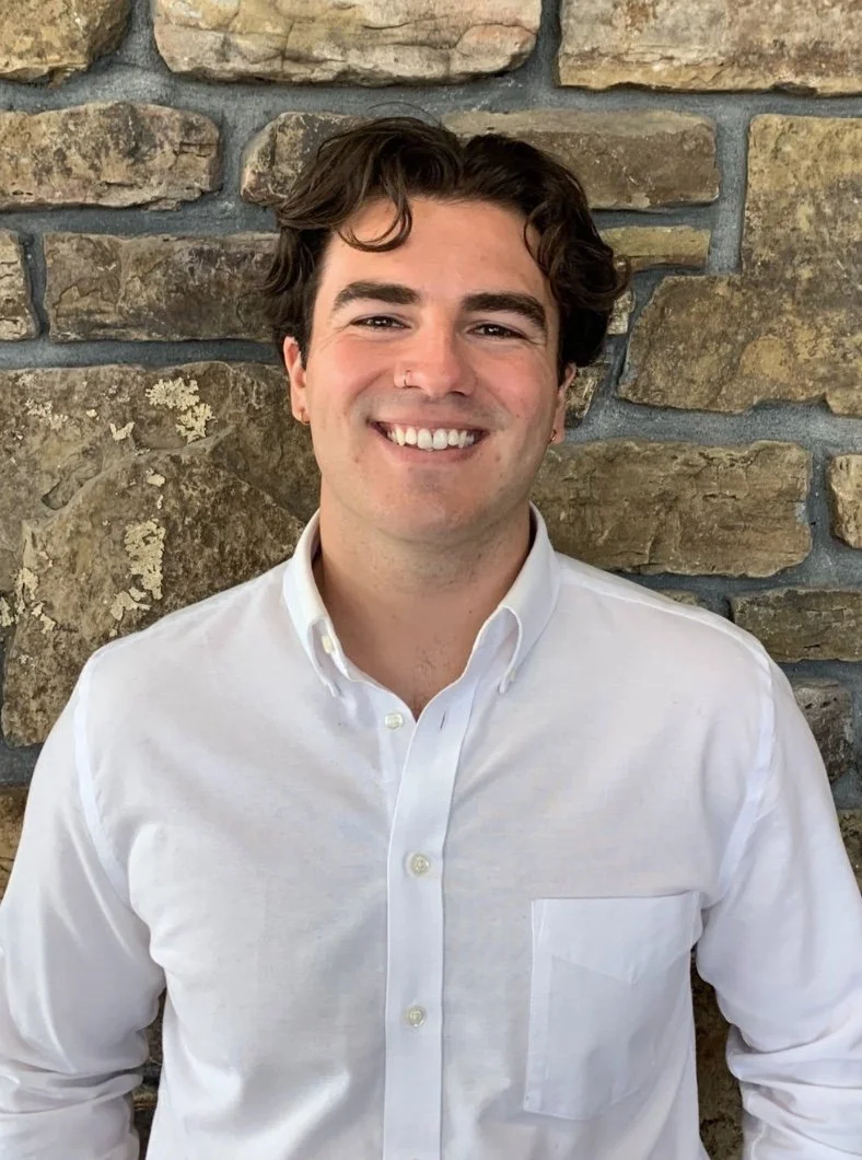 A smiling young man with dark curly hair and a nose piercing, wearing a white button-up shirt, standing against a stone wall background.