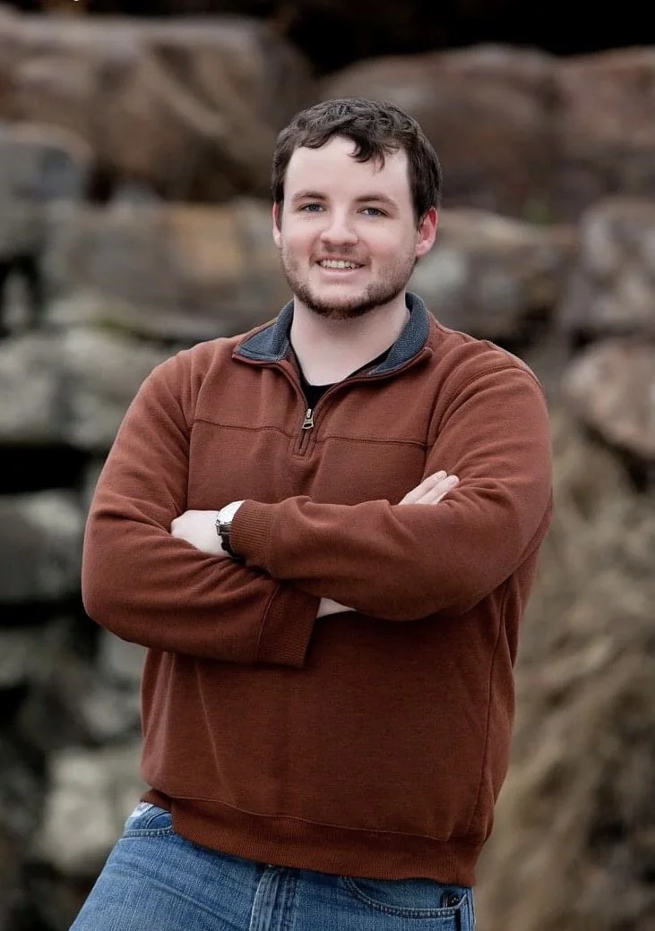 A young man with short dark hair and beard smiling, standing outdoors with arms crossed, wearing a brown pullover and blue jeans, with rocks in the background.