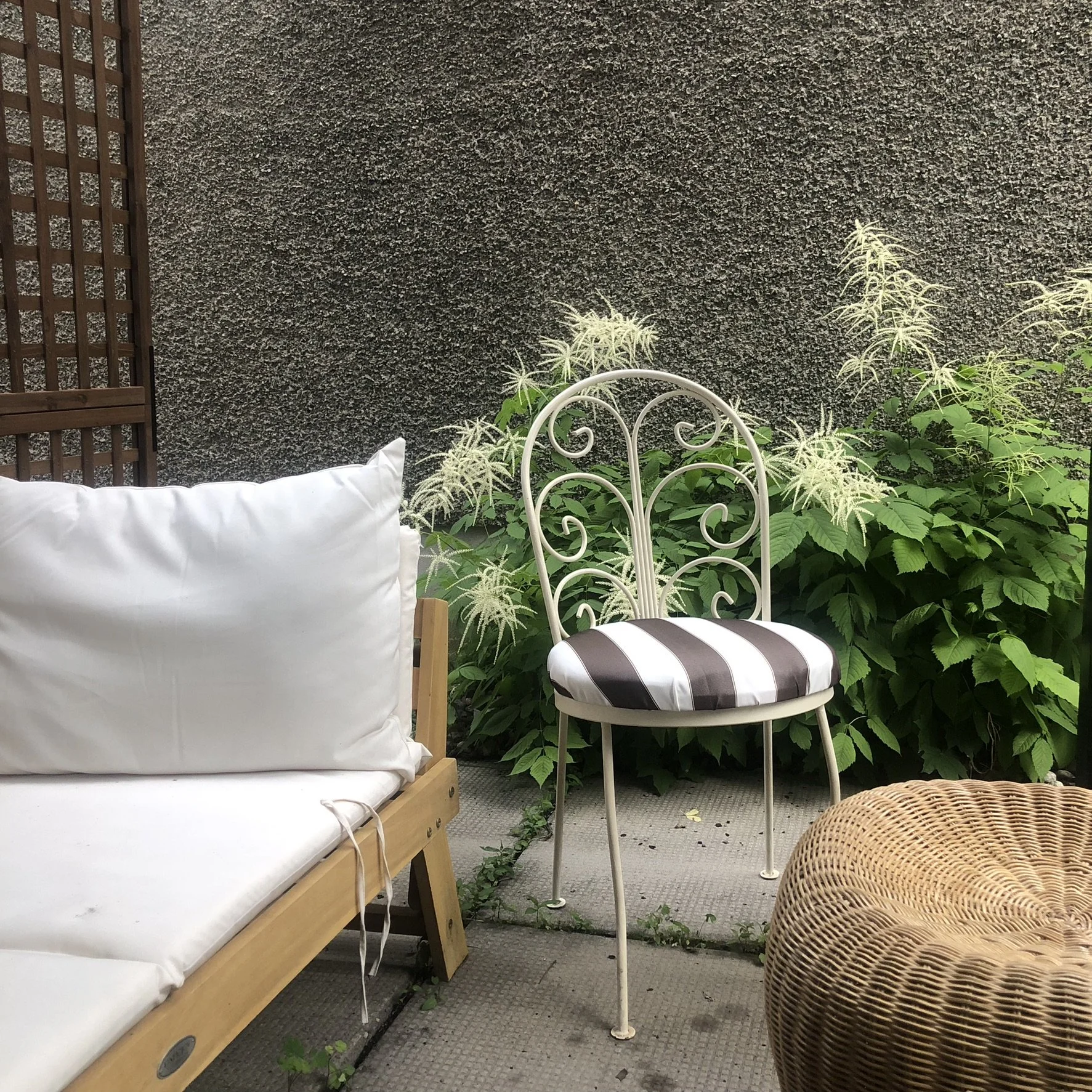 Outdoor patio area with white wicker chair, striped cushion, white metal chair, wicker ottoman, and a bed with white linens, surrounded by lush green plants with white flowers and a textured gray wall in the background.