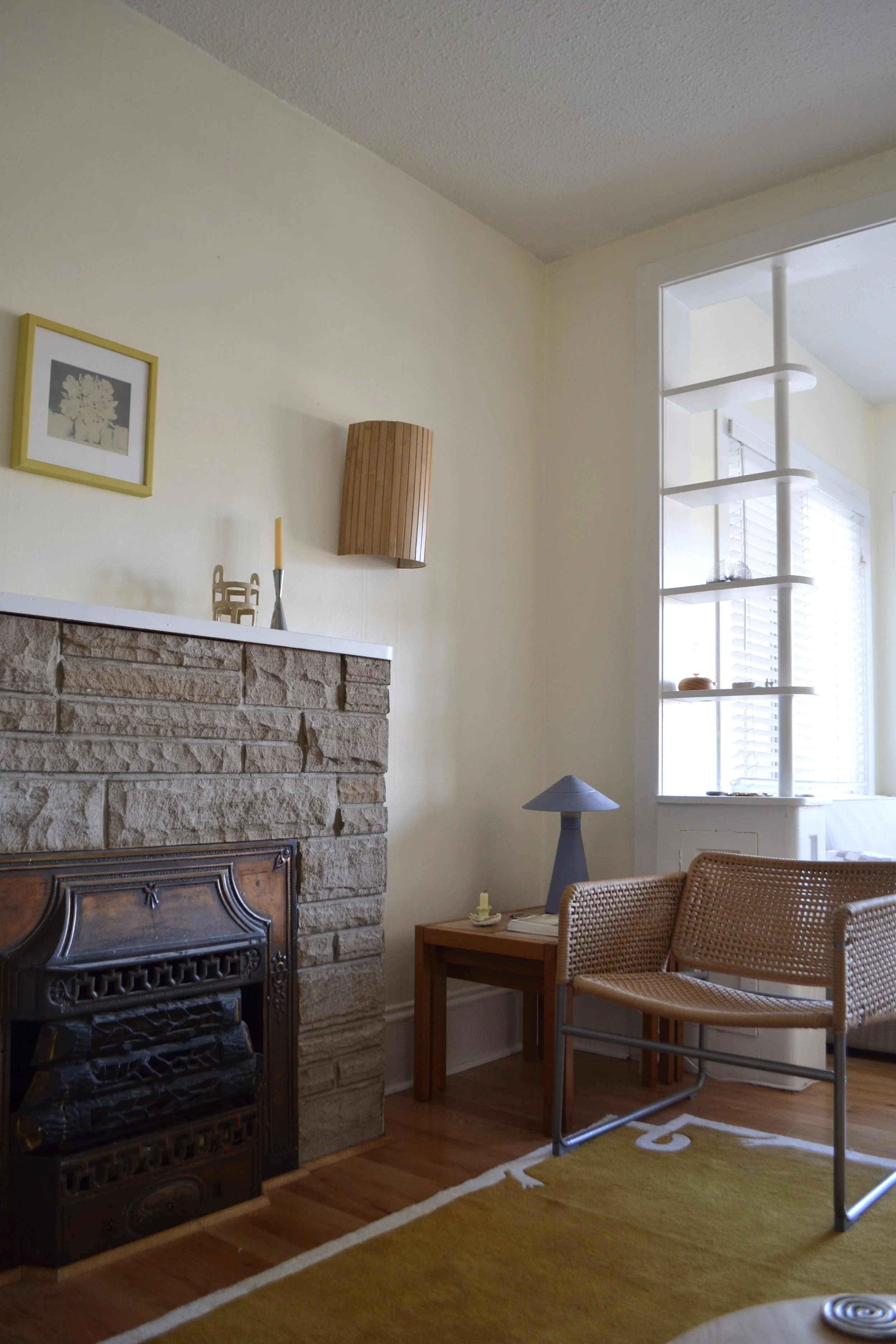 Living room corner with a stone fireplace, a small wooden side table, a rattan chair, and a large window with shutters. Decor includes a table lamp, picture frame, and wall-mounted light fixture.