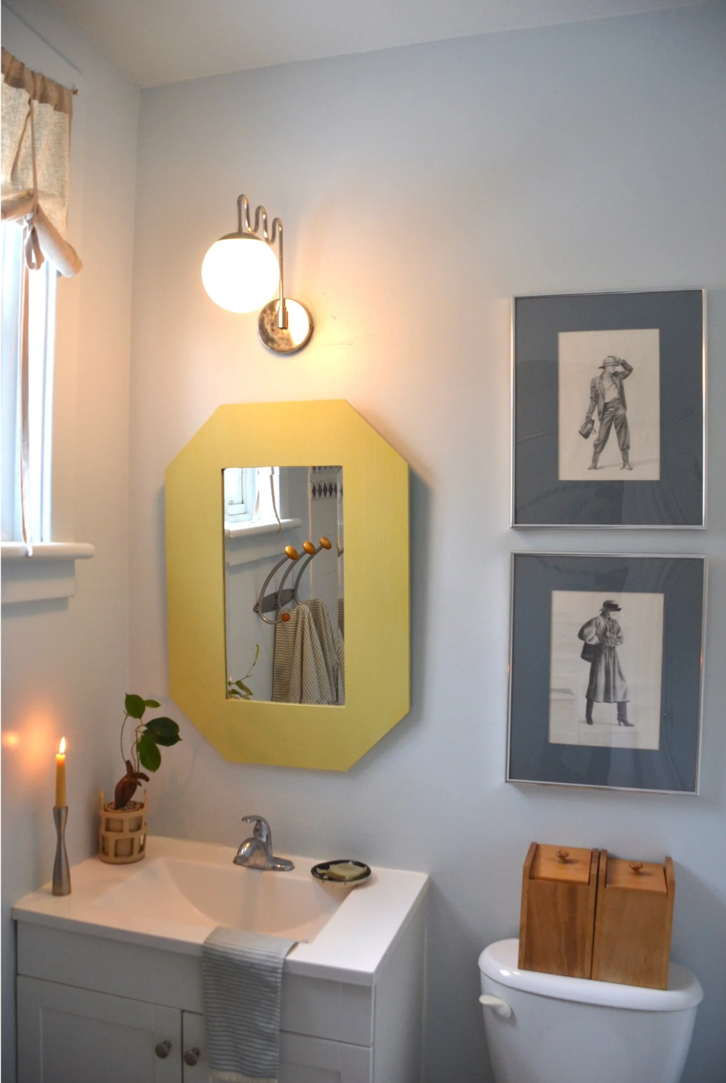 A bathroom with white walls, a yellow-framed mirror, framed black and white photos of women in stylish outfits, a white vanity with a small plant, a candle, and a soap dish, a wooden cabinet on a toilet, and a metal wall sconce with a globe light.