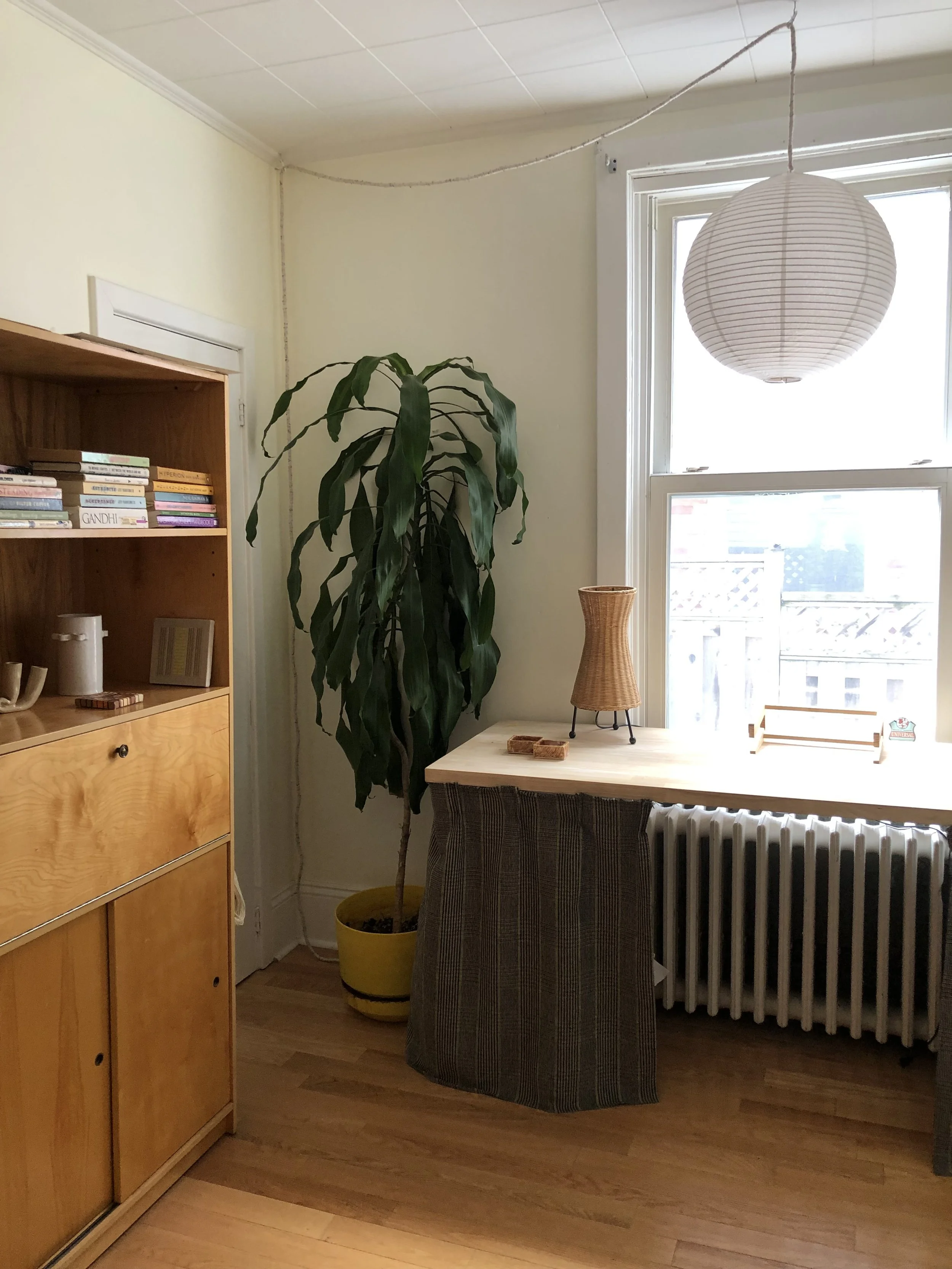 A room with a yellow potted plant next to a wooden cabinet, a table with a wicker lamp and a tray, a window with a white radiator underneath, a white paper lantern hanging from the ceiling, and books on the cabinet.