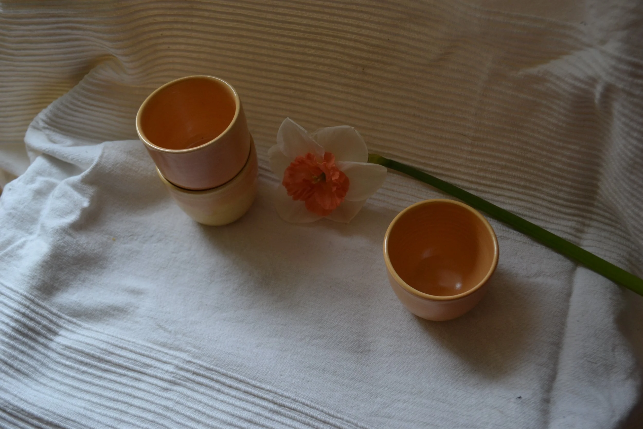 A white textured cloth with a pink ceramic cup, a white flower with orange edges, and an orange cup, all arranged on it.