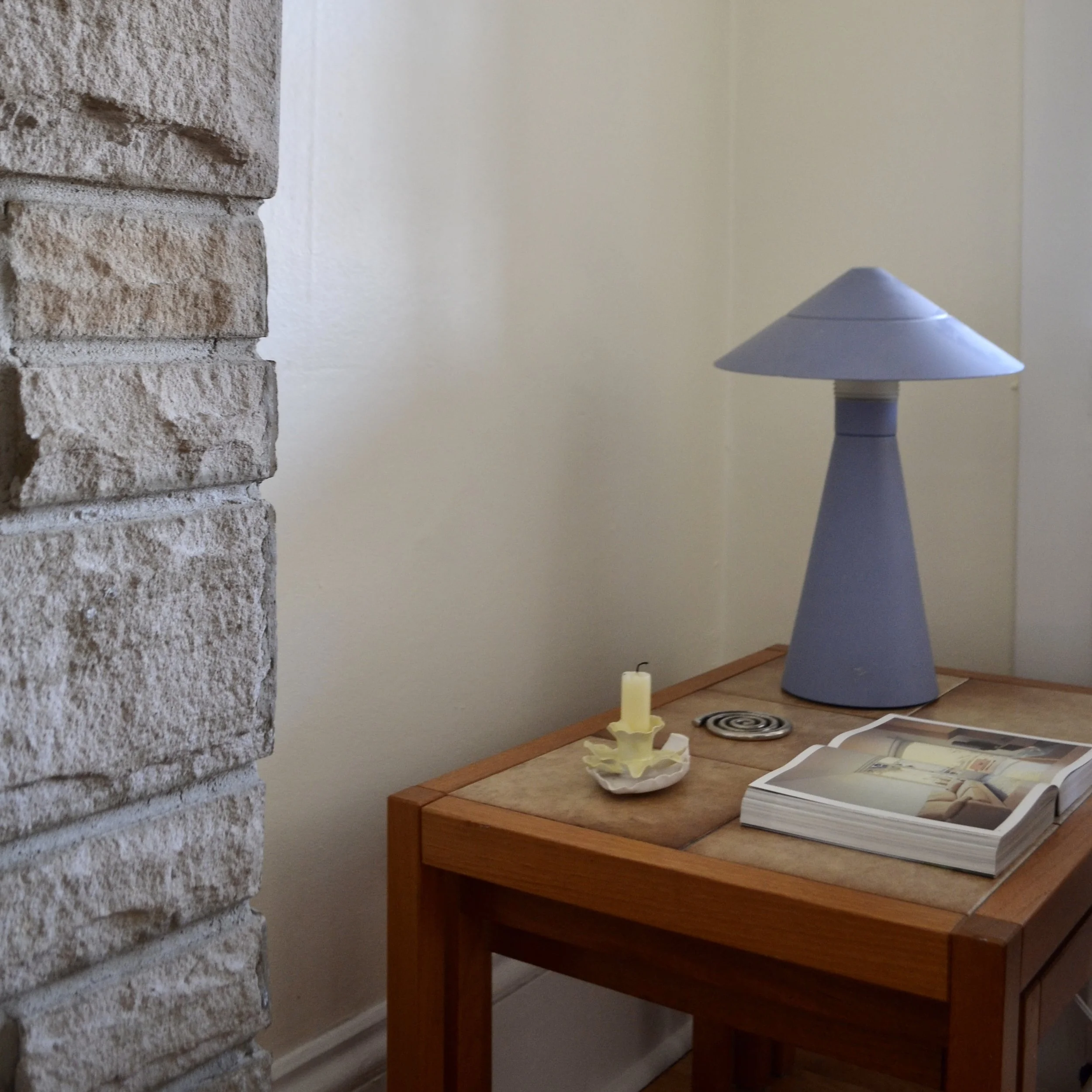 Interior scene with a stone wall on the left, a wooden table with magazines, a yellow candle, and a small sculpture, a blue lava lamp, and a cream-colored wall in the background.