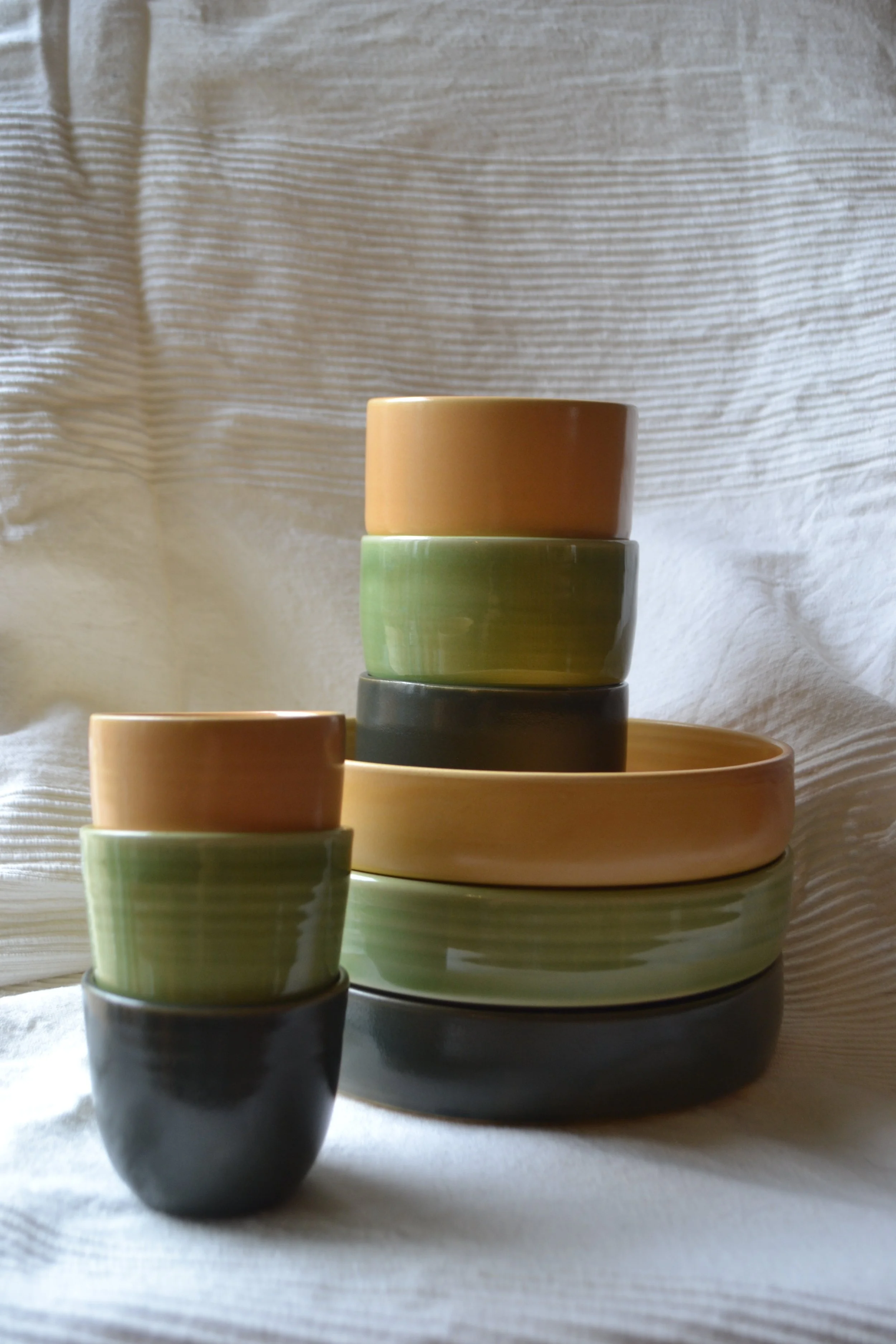 Stacked ceramic bowls in beige, green, and black colors on a table with a backdrop of textured white fabric.