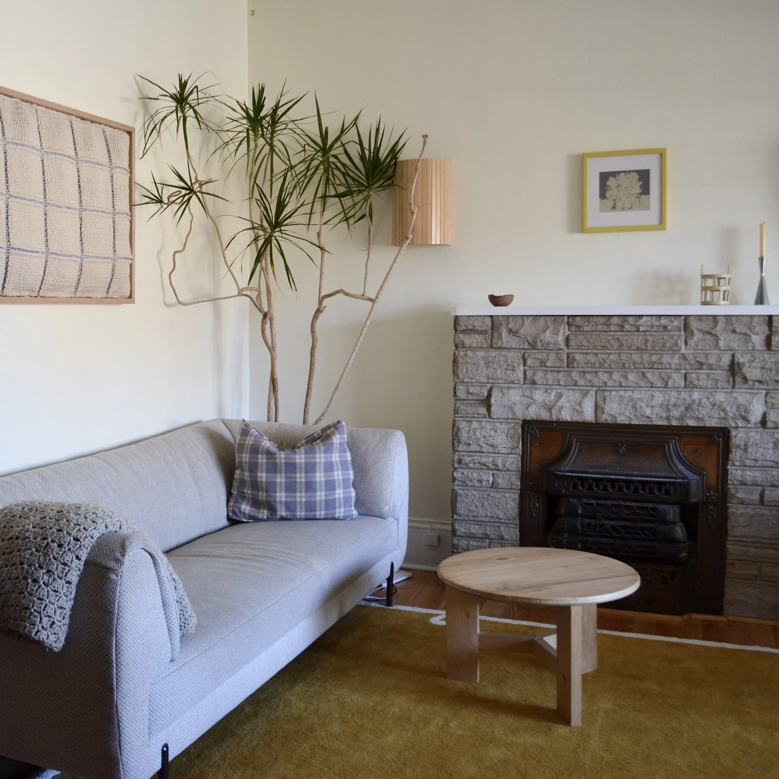 Living room with a beige sofa, a plaid pillow, a round wooden coffee table, a fireplace with a stone surround, a tall houseplant next to a wall decorated with a framed artwork, a candle holder, and a lamp.