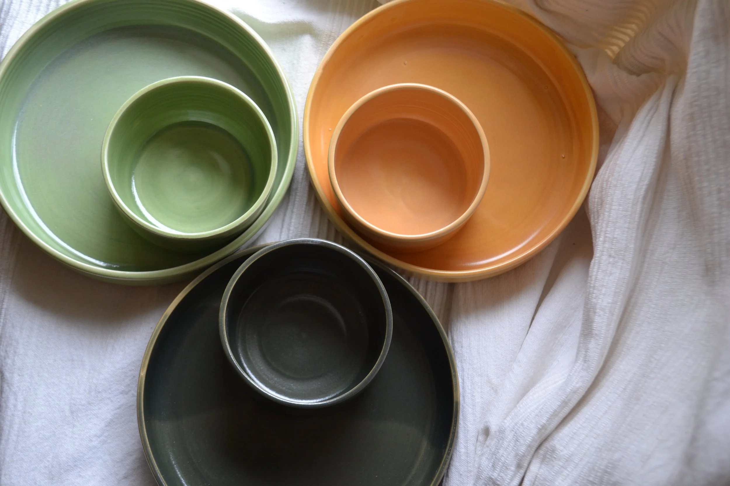 A collection of nested ceramic bowls and containers in green, yellow, black, and orange on a white surface with a cloth nearby.