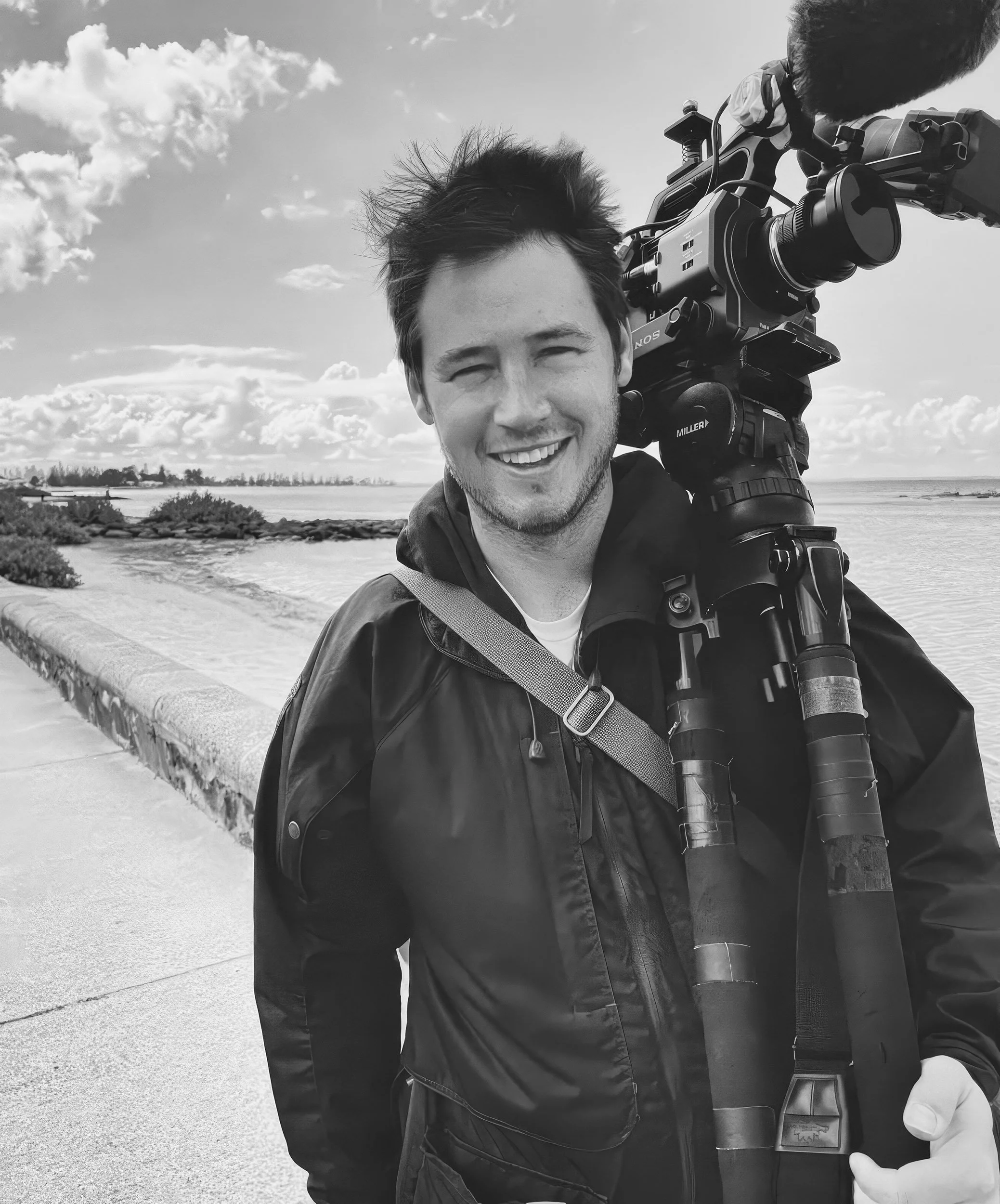 A smiling man with dark hair, holding a professional video camera on his shoulder, standing outdoors near a body of water with a cloudy sky overhead.