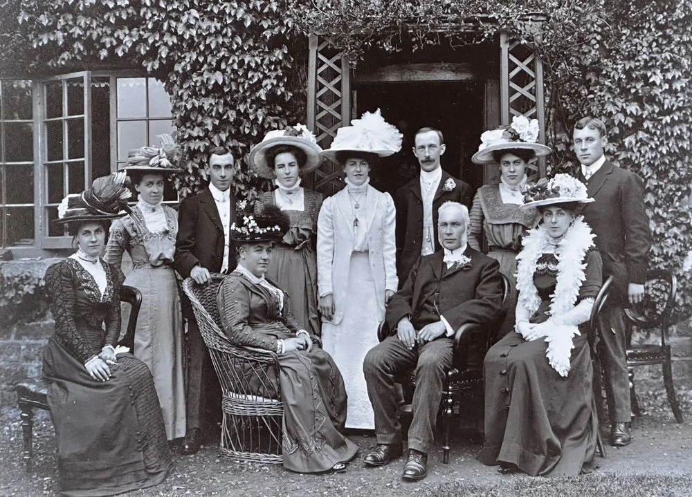 A black-and-white photograph of ten people, four men and six women, posing outdoors near a house with ivy-covered walls and large windows. The women are dressed in Victorian-era attire with large hats adorned with flowers and feathers. The men are dr