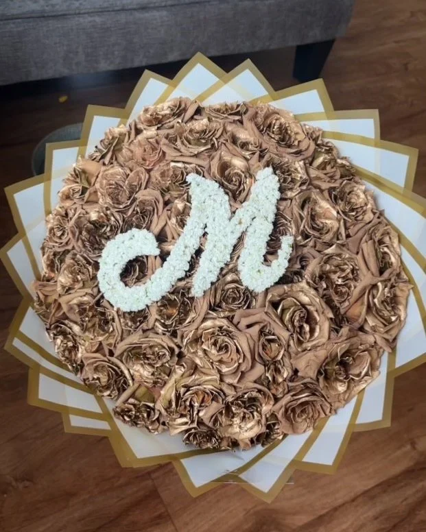 A round bouquet of gold-colored roses with a white cursive letter "M" in the center, surrounded by a decorative paper or cardboard border.