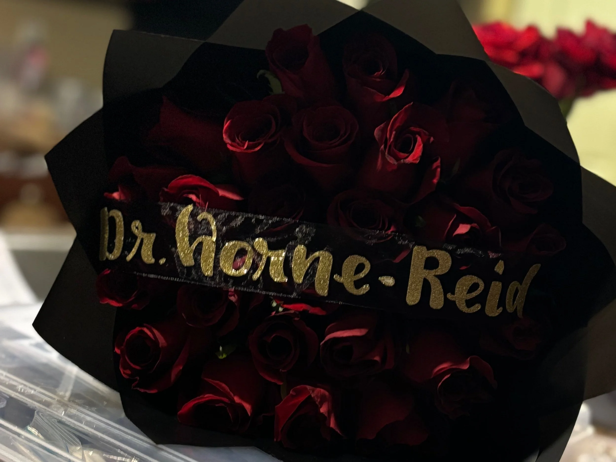 A bouquet of red roses wrapped in black paper with a black ribbon that has gold lettering saying, 'Dr. Horne-Reid.'