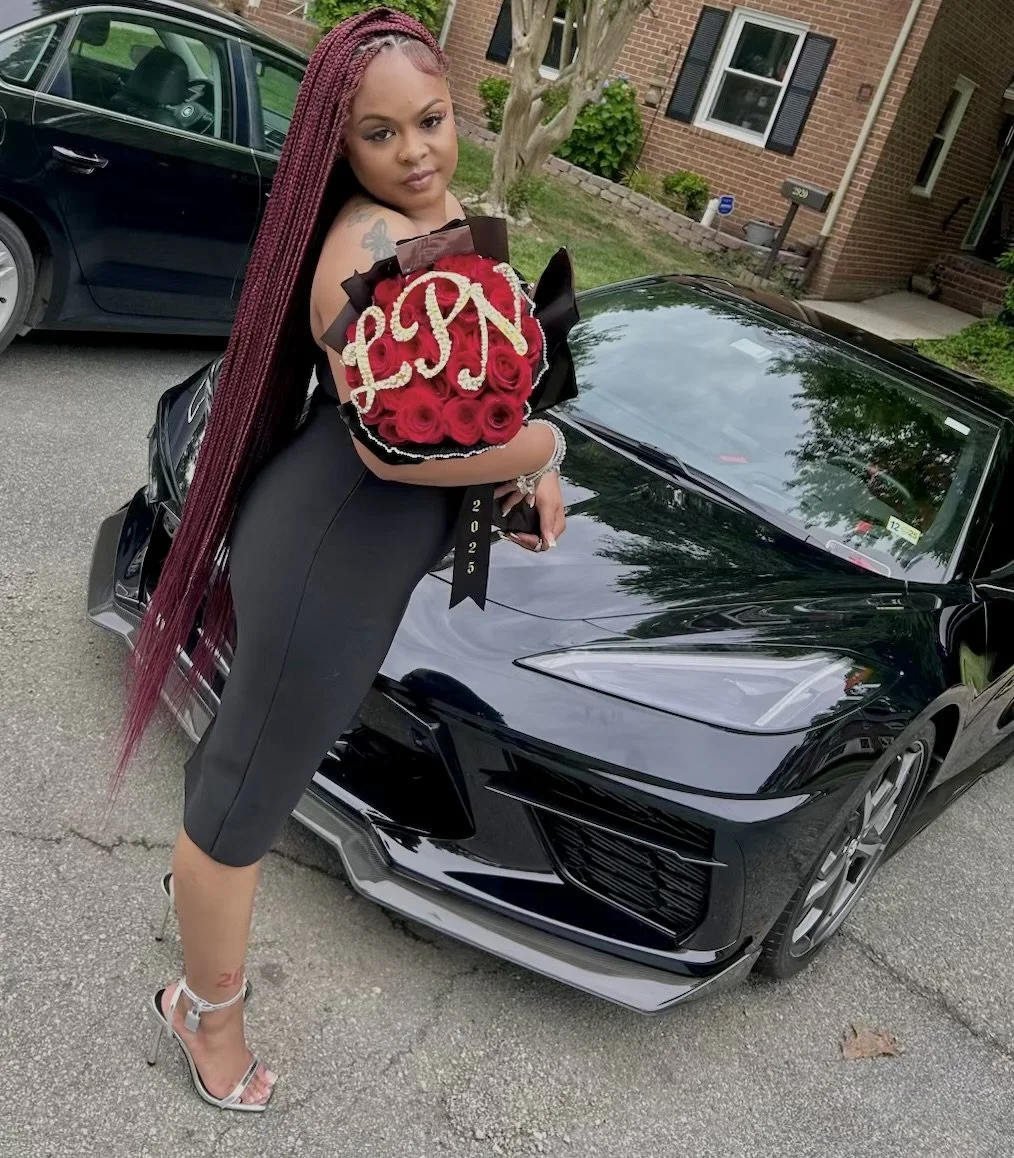 A woman with long burgundy braided hair holding a bouquet of red roses with the initials 'LCN' in white roses, standing next to a black sports car on a residential street.