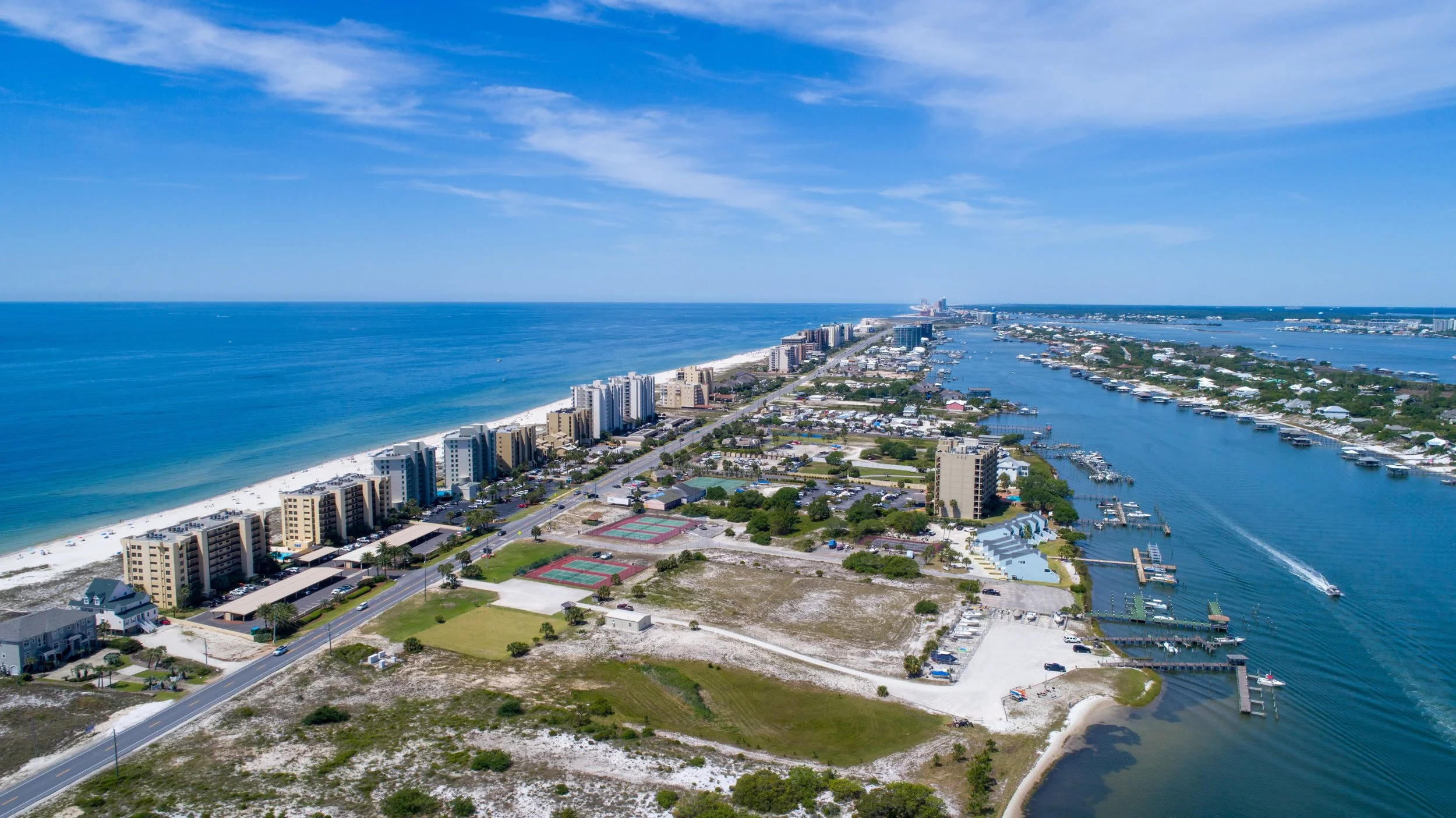 Aerial view of a coastal city with beachfront buildings, tennis courts, and a marina with boats along a river, under a partly cloudy sky.