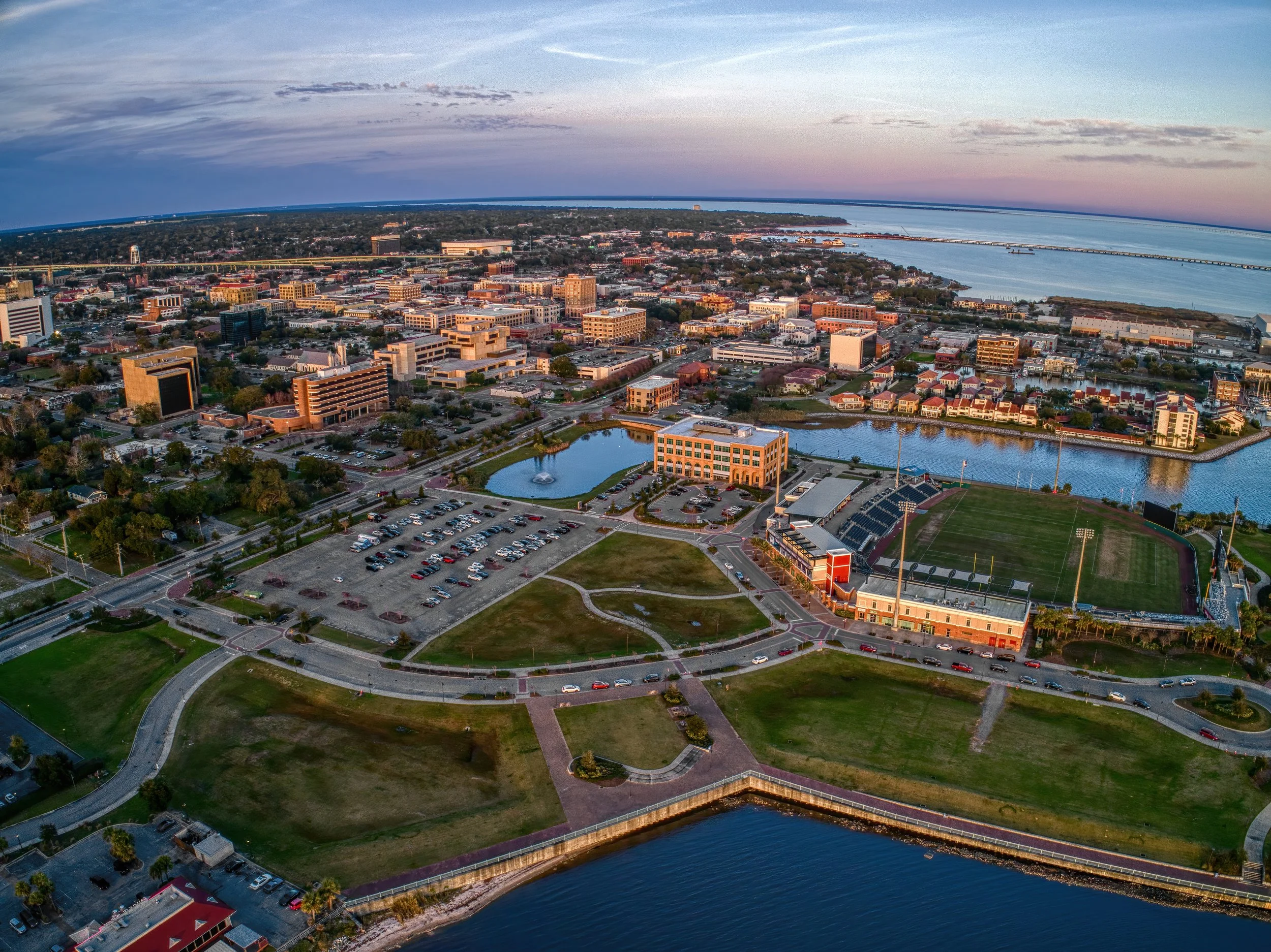 Aerial view of a cityscape at sunset showing a sports stadium with a football field, a large parking lot, water bodies, and numerous buildings near the coast.