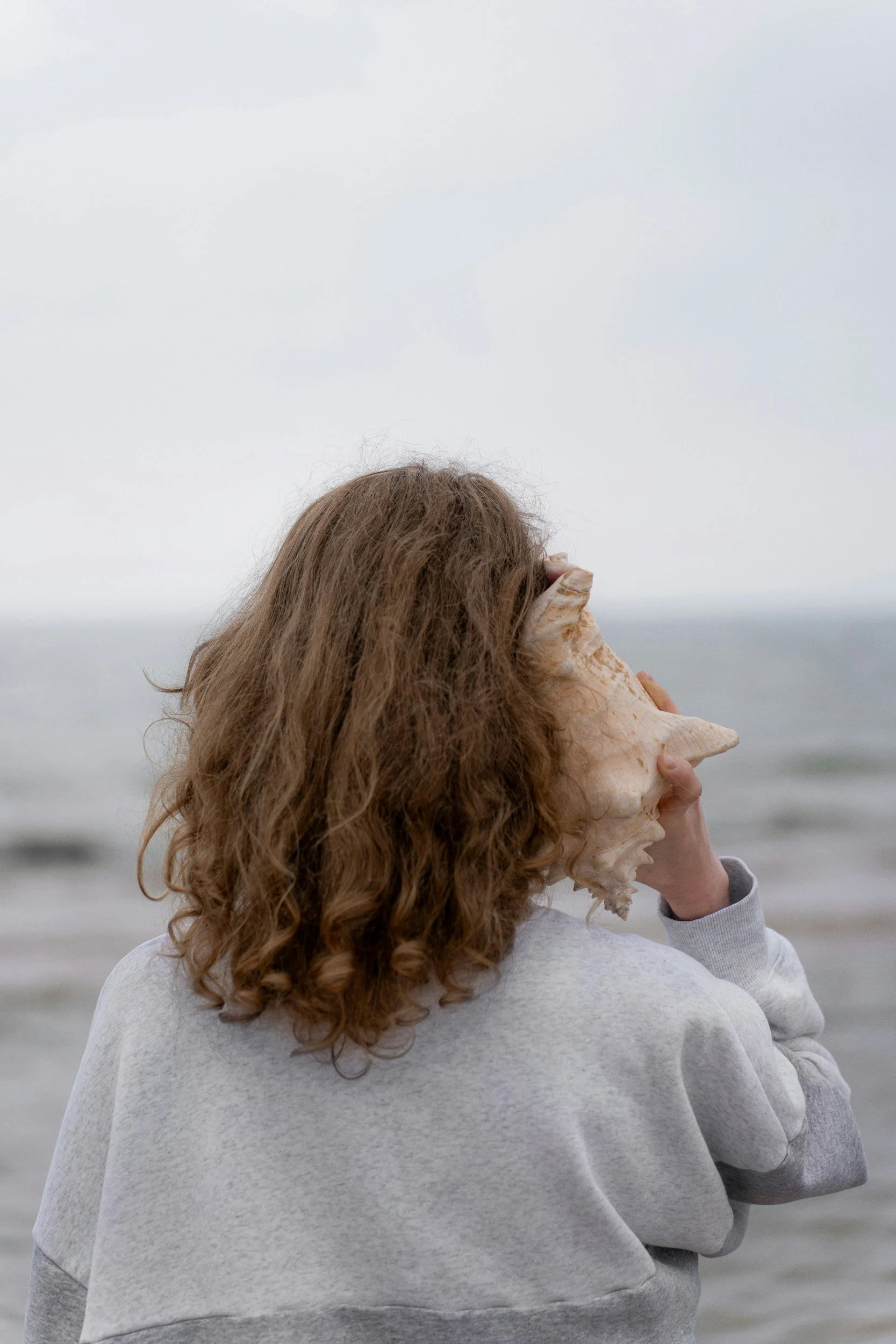 A person with curly brown hair wearing a grey hoodie holding a large seashell up to the side of their face at the beach.