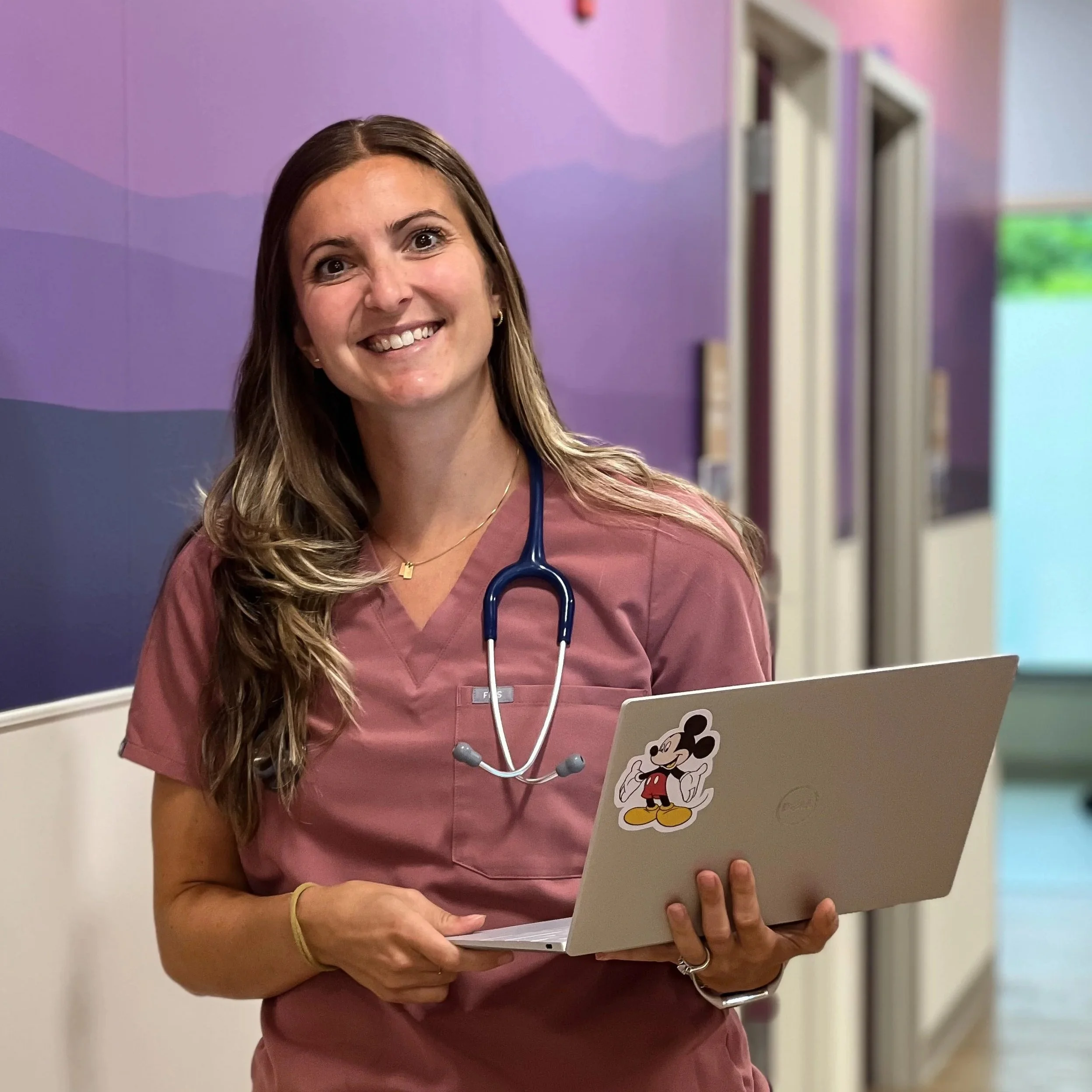 A smiling female healthcare worker in pink scrubs holding a laptop with a Mickey Mouse sticker, wearing a stethoscope around her neck, standing in a colorful medical facility hallway.