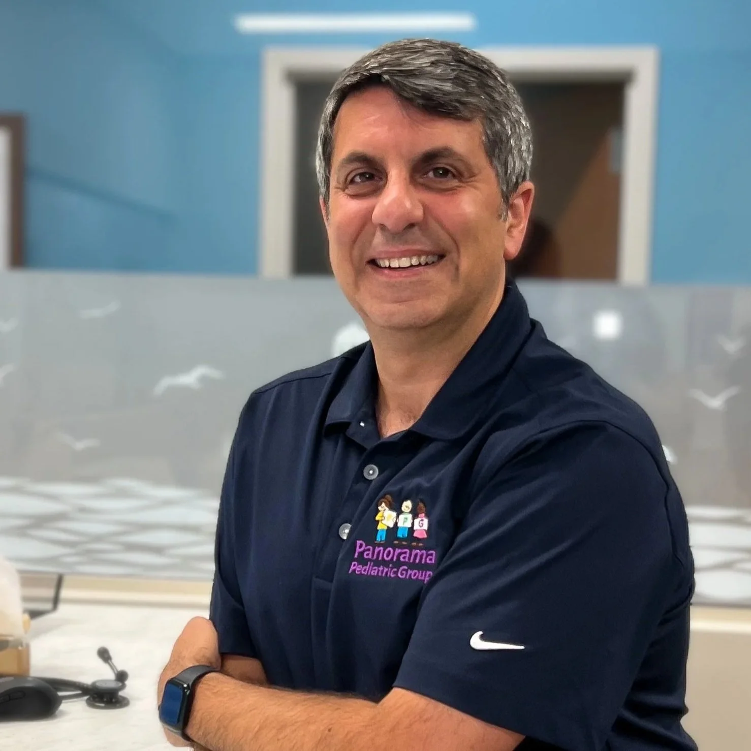 A smiling man with gray hair in a navy blue polo shirt with a logo that reads "Panorama Pediatric Group" stands with his arms crossed, wearing a smartwatch, background features a blue wall and a glass partition.