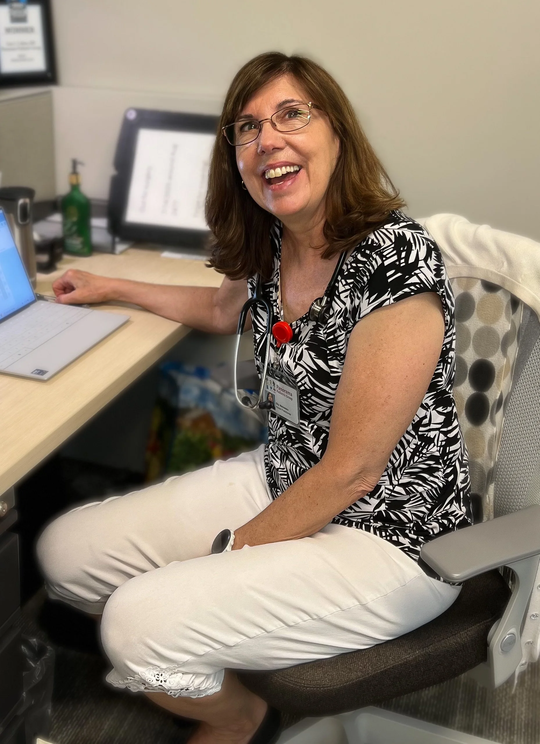 A woman with glasses, smiling, wearing a black and white patterned top, cream-colored pants, and a stethoscope around her neck, sitting at a desk in an office or medical setting.