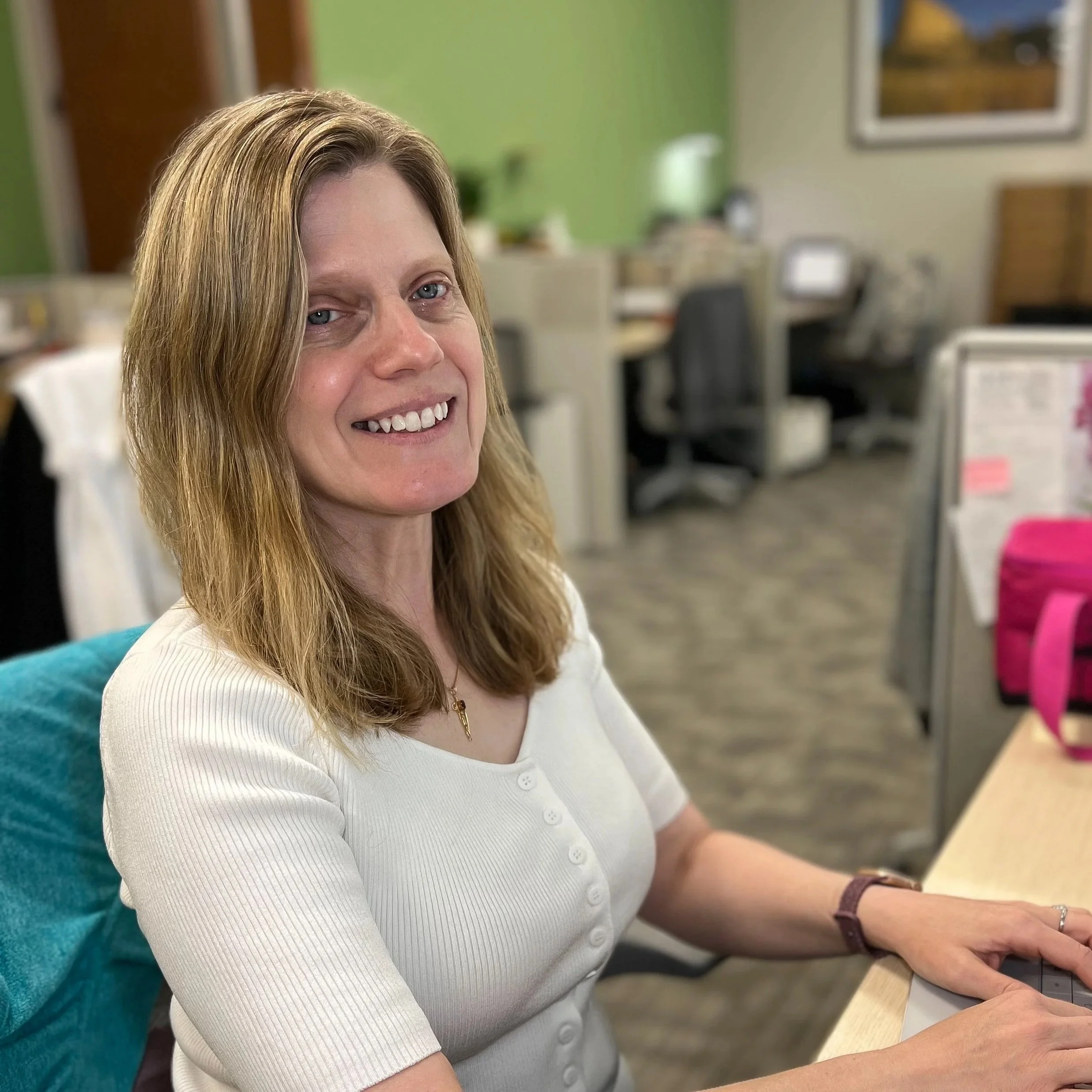 A woman with shoulder-length light brown hair, smiling while working at a desk in an office.