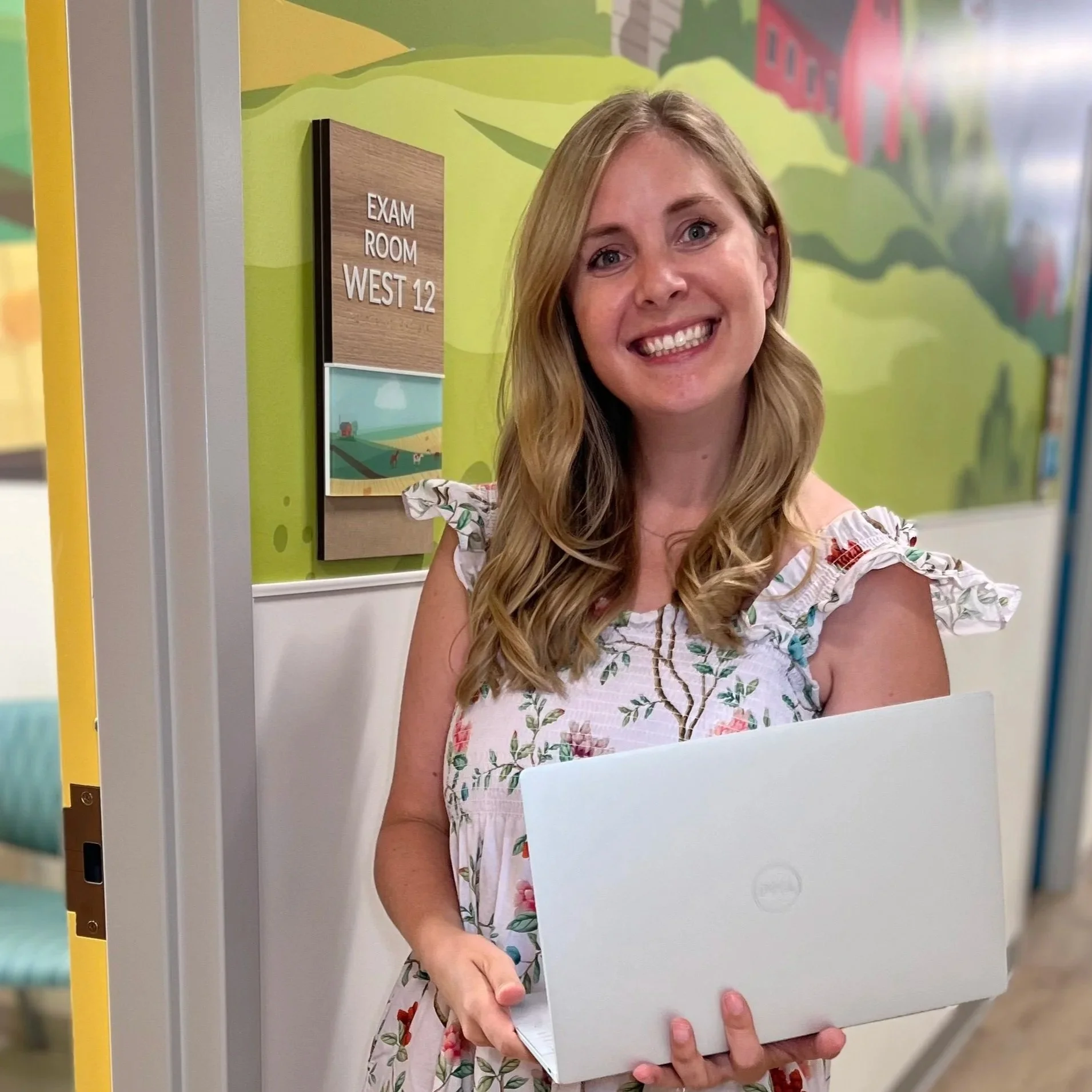 A woman with long wavy blonde hair smiling while holding a closed white laptop in front of her. She is wearing a white dress with a floral pattern. The setting is indoors near a door that says "Exam Room West 12" on a wooden sign, with a colorful, cartoon-style mural in the background.