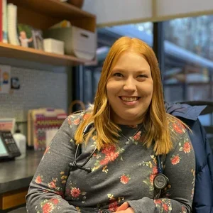 A woman with red hair smiling in a medical office, wearing a floral sweater and stethoscope.