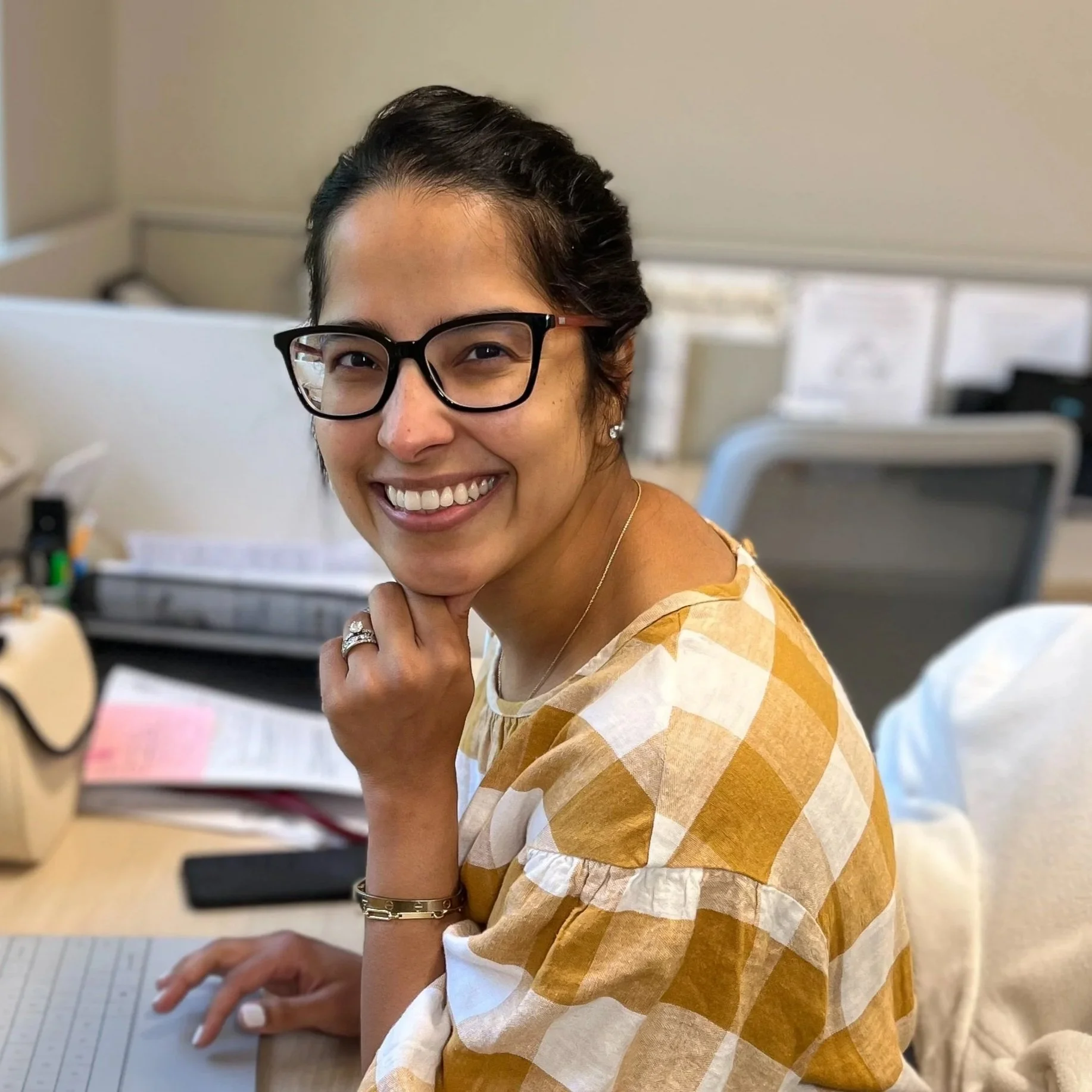 A woman with dark hair tied back, wearing glasses, a gold ring, bracelet, and necklace, smiling while sitting at a desk with various office supplies and papers in the background.