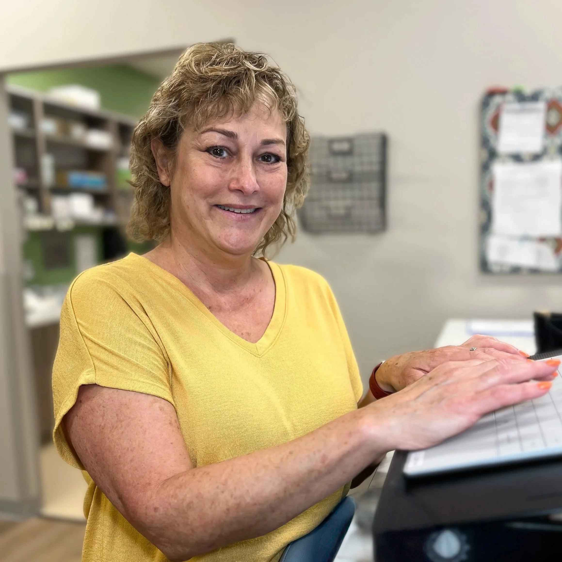 A woman wearing a yellow shirt smiling while working on a tablet in an office setting.