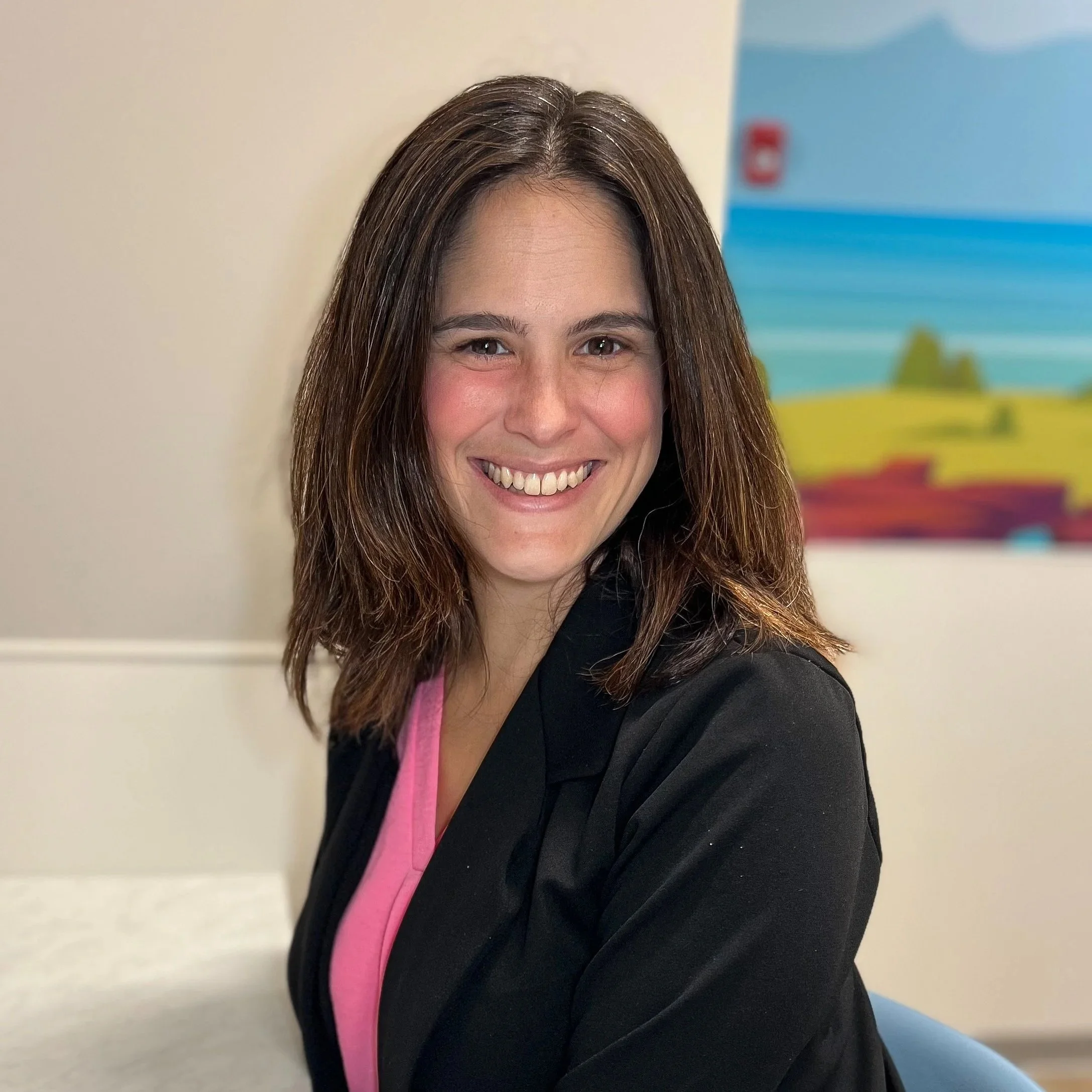 A woman with shoulder-length brown hair smiling at the camera, wearing a black jacket over a pink shirt, with a colorful landscape painting in the background.