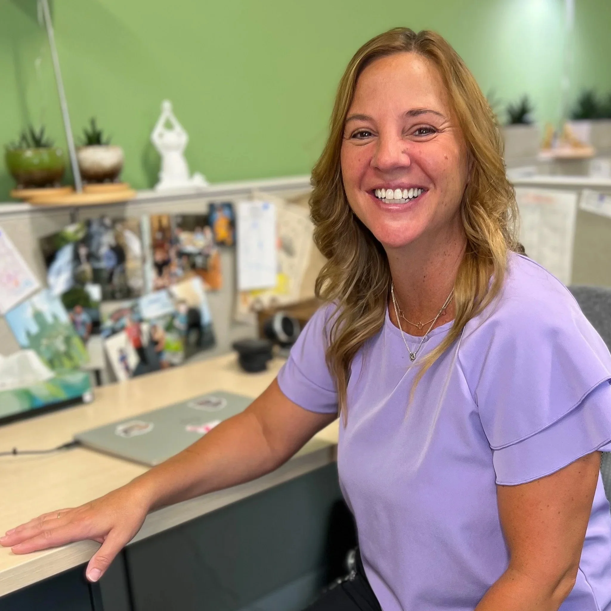 A woman with shoulder-length reddish hair sitting at a desk in an office cubicle, smiling at the camera. She is wearing a light purple blouse and jewelry, with a green wall and personal items in the background.