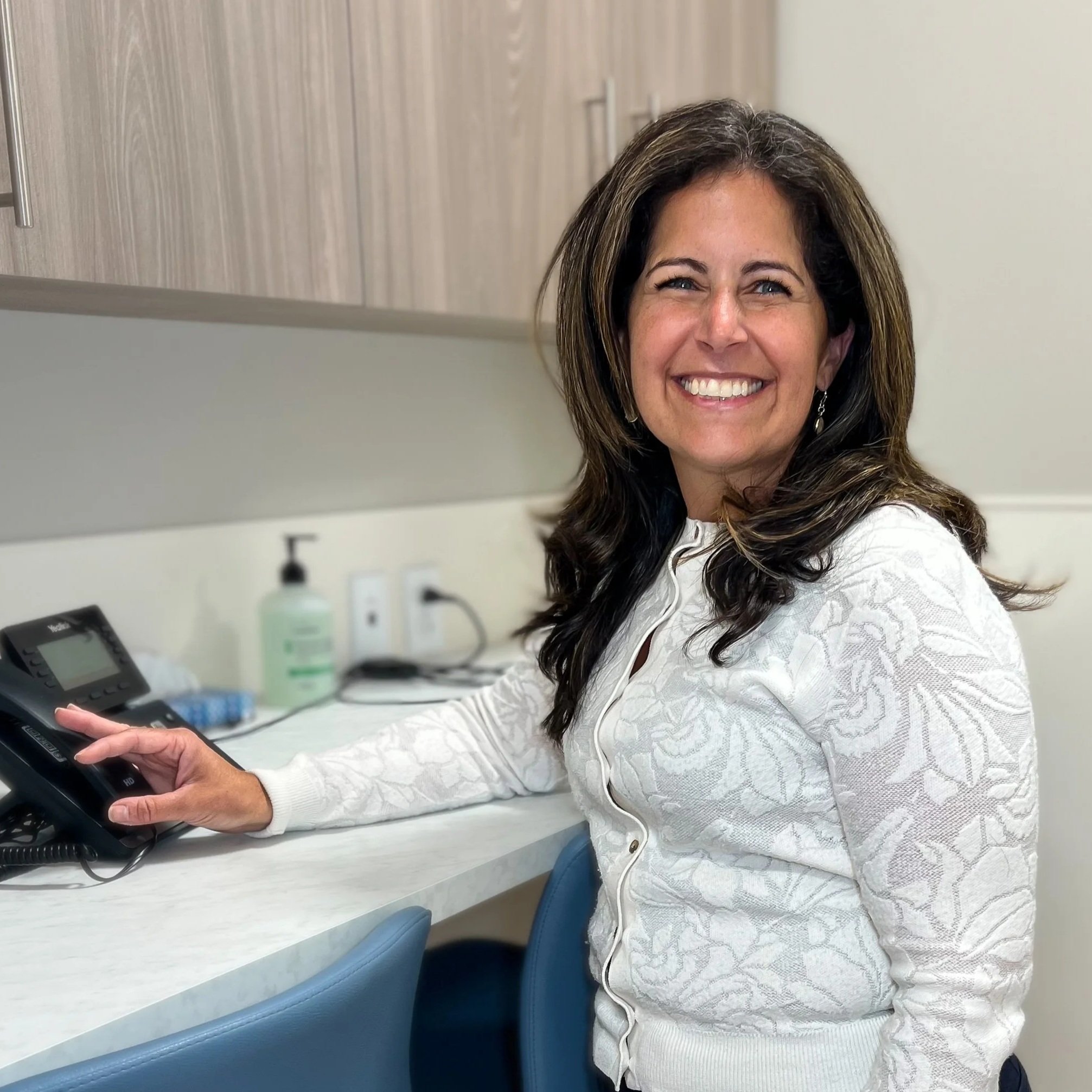 A woman with brown hair and blue eyes, smiling, sitting in a medical office, holding a phone on a countertop.