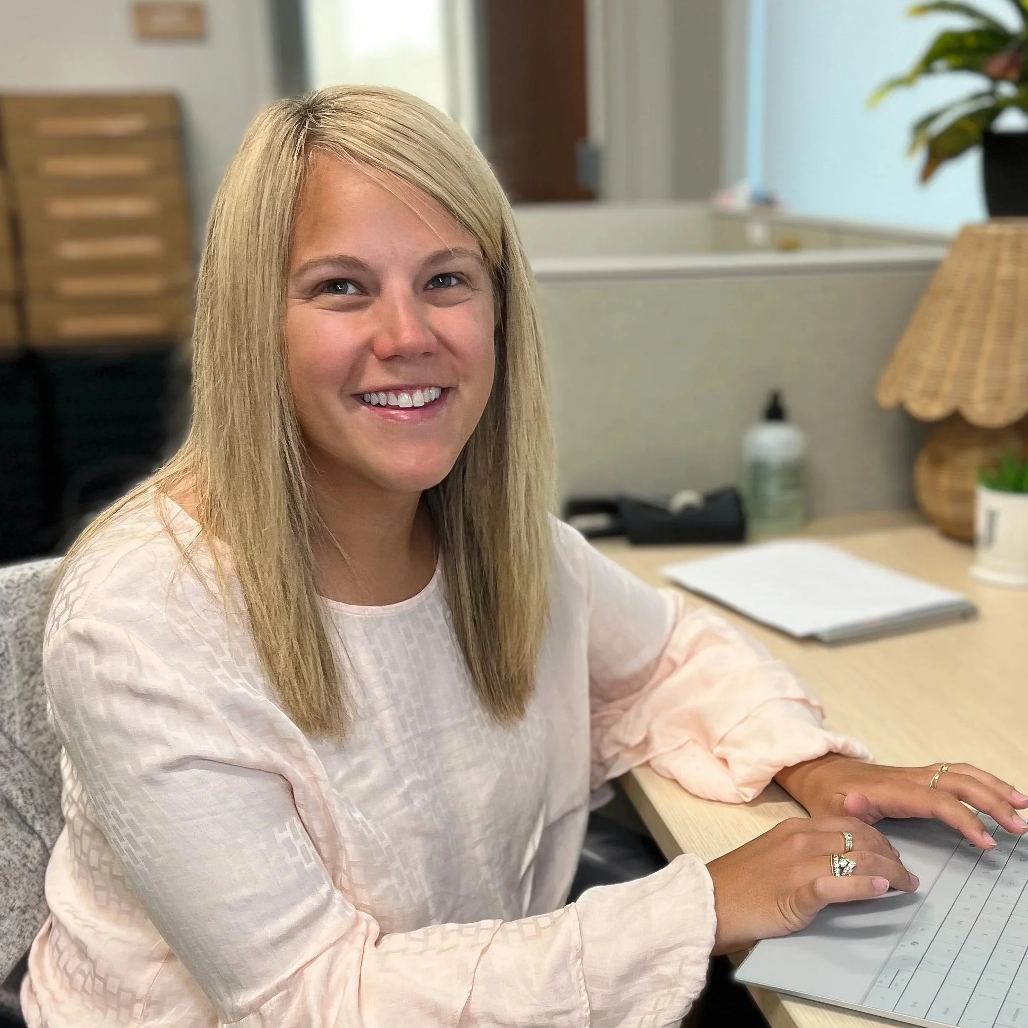 A smiling woman sitting at a desk with office supplies, documents, and a computer, in a professional office setting.