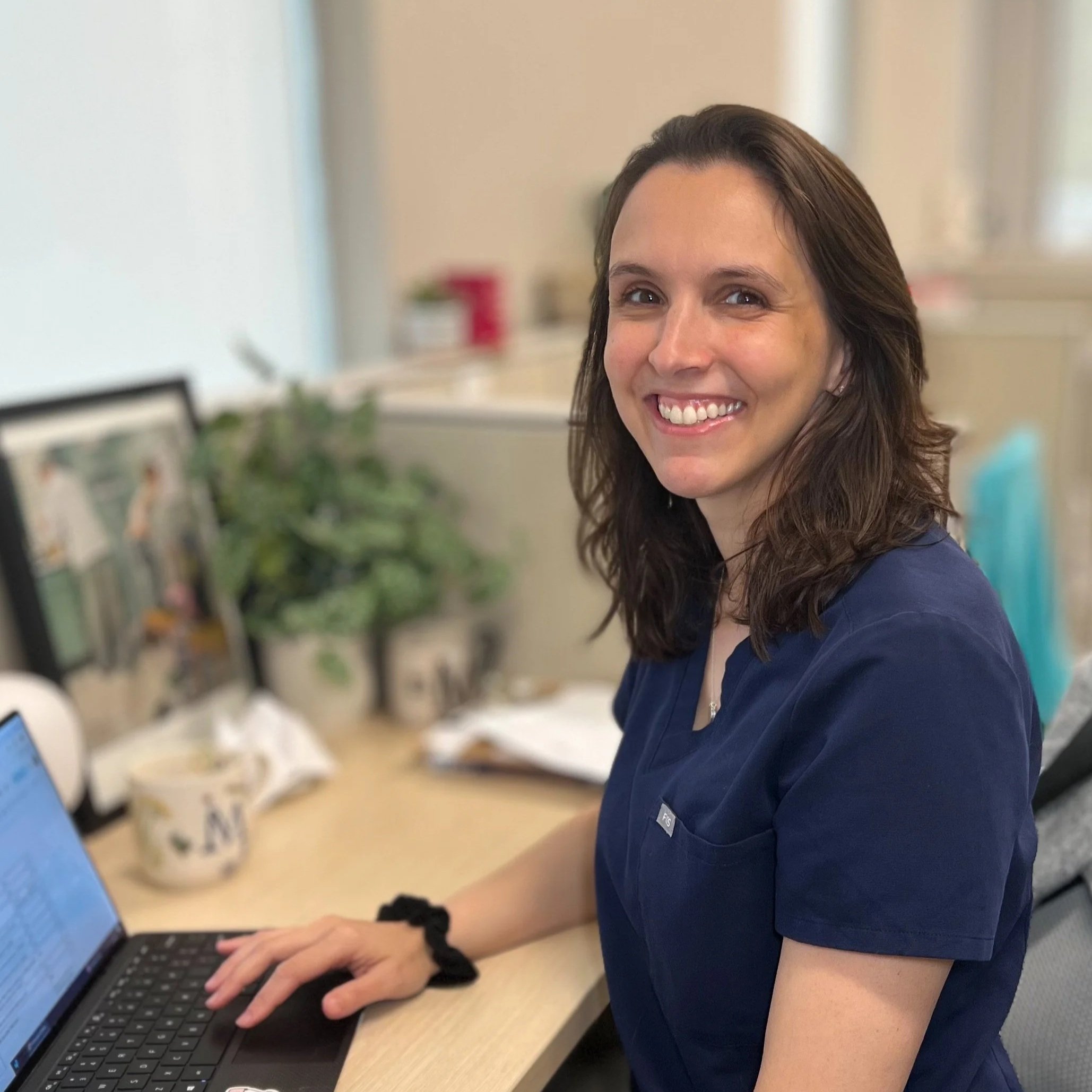 A woman in navy scrubs sitting at a desk with a laptop, smiling at the camera in an office setting.