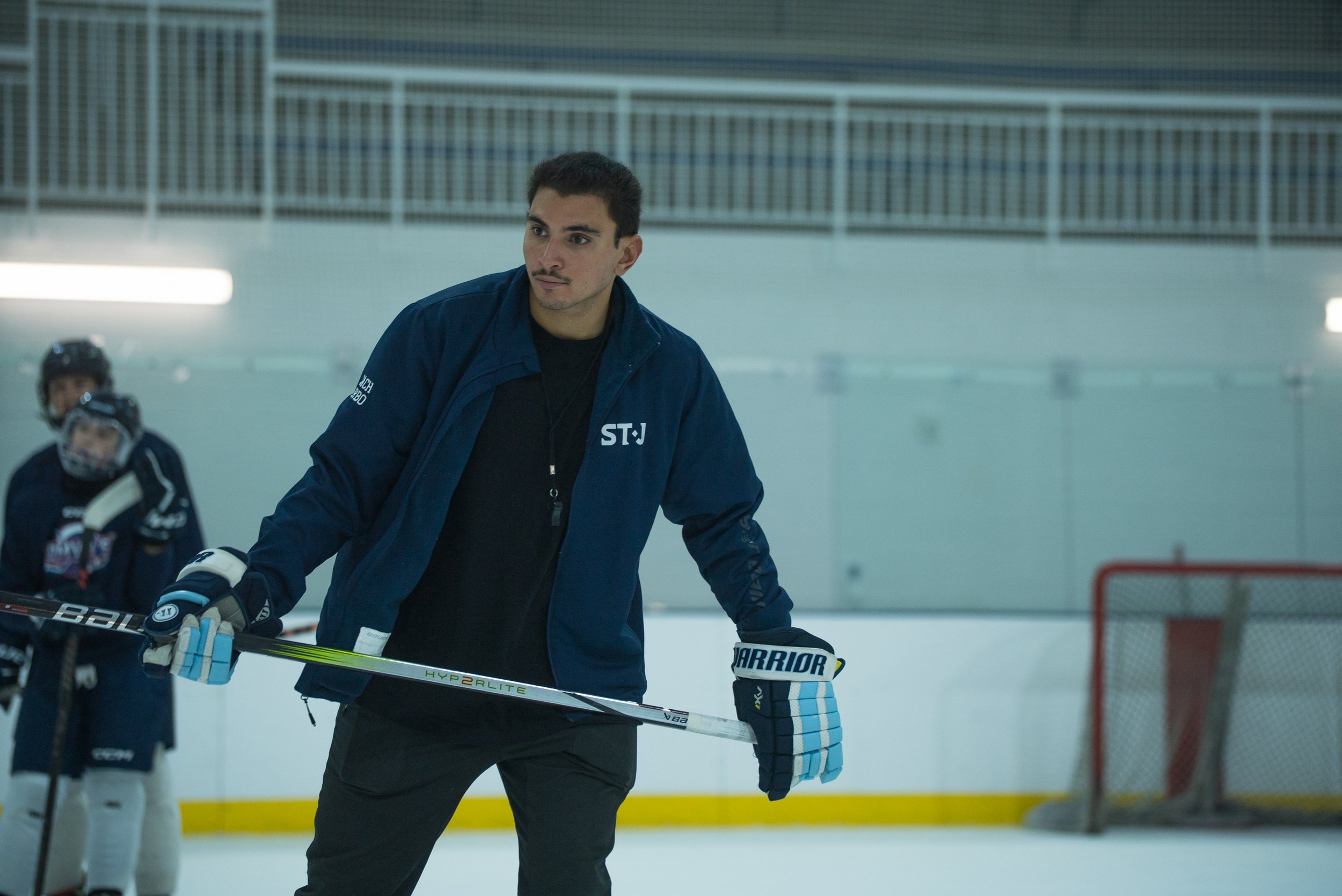 A young male hockey coach standing on the ice rink during practice, holding a hockey stick, with young players and a goal net in the background.