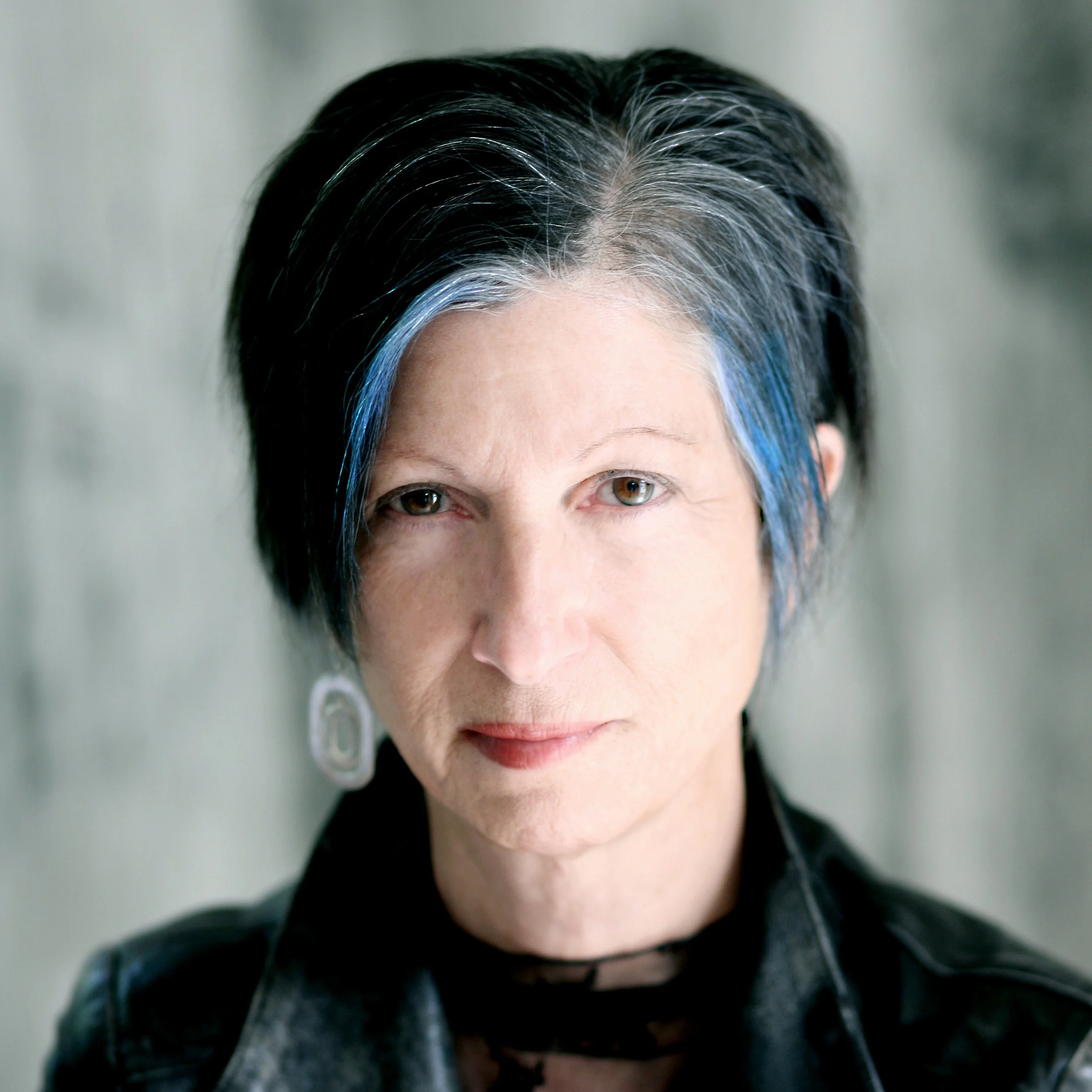 A close-up portrait of a woman with short black hair with blue streaks, wearing earrings and a black leather jacket, looking directly at the camera.