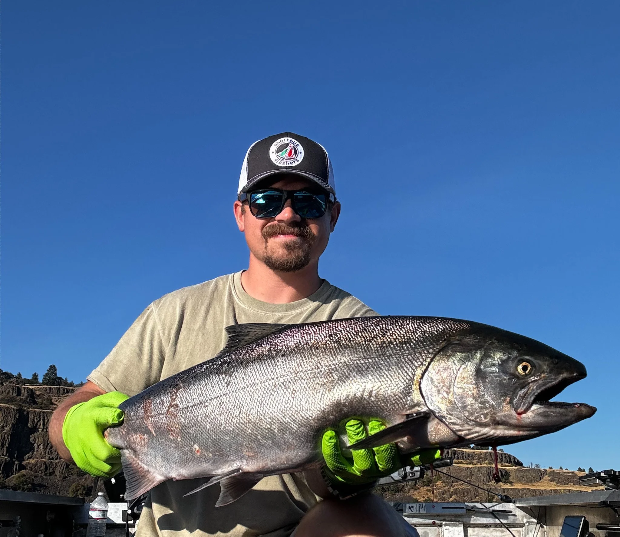 Man wearing sunglasses, a black and white cap, and neon green gloves holding a large fish against a clear blue sky with hills in the background.