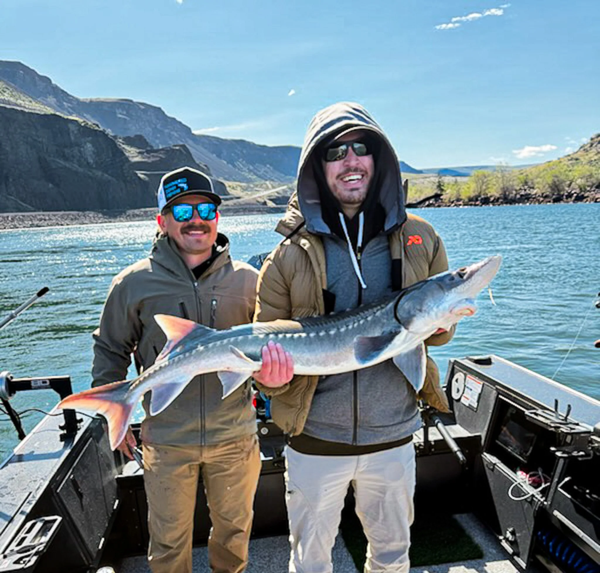 Two men on a boat holding a large fish with mountains, water, and a blue sky in the background.