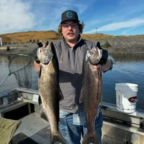 Young man holding two large fish, one in each hand, on a boat by the water during daytime.